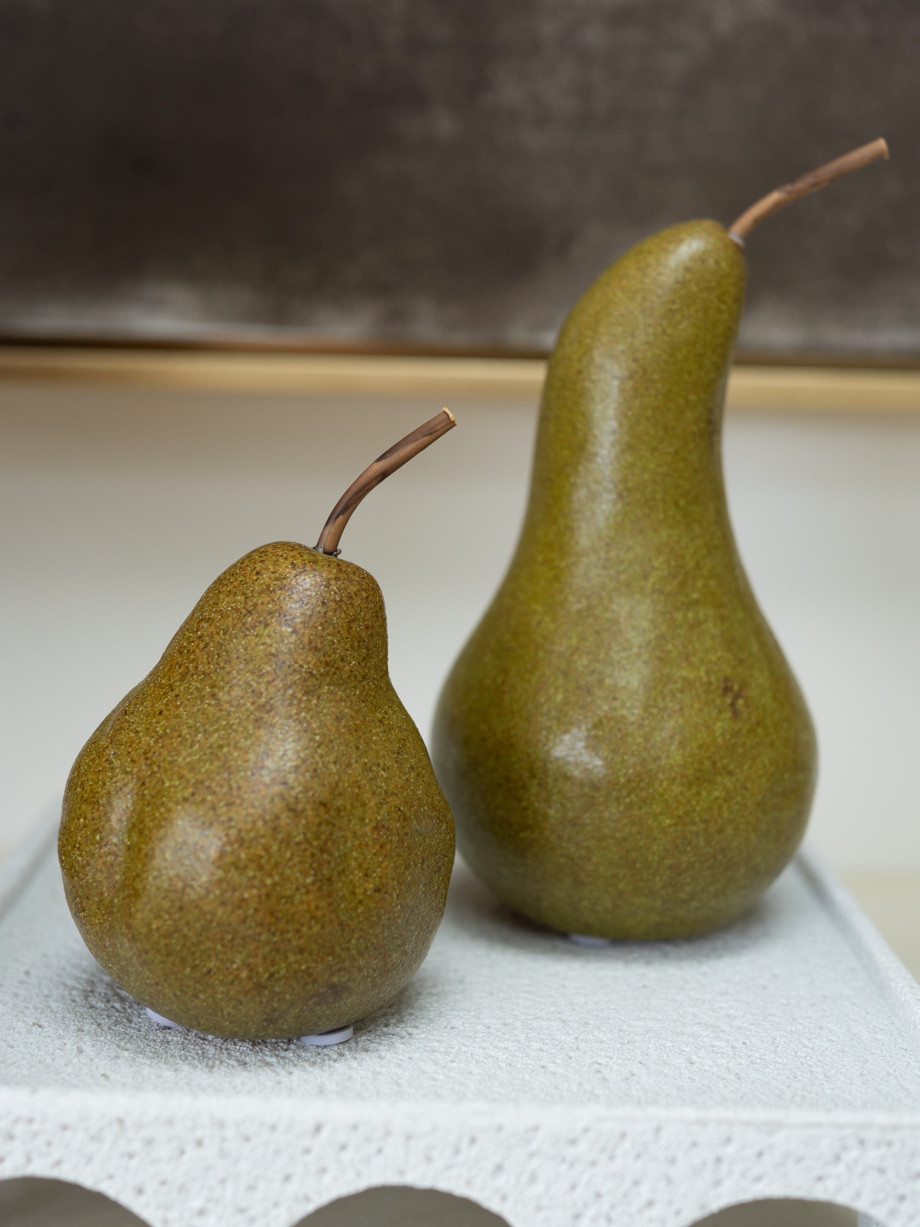 Two whole pears with brown stems, one smaller and one larger, standing on a white textured surface against a neutral background.