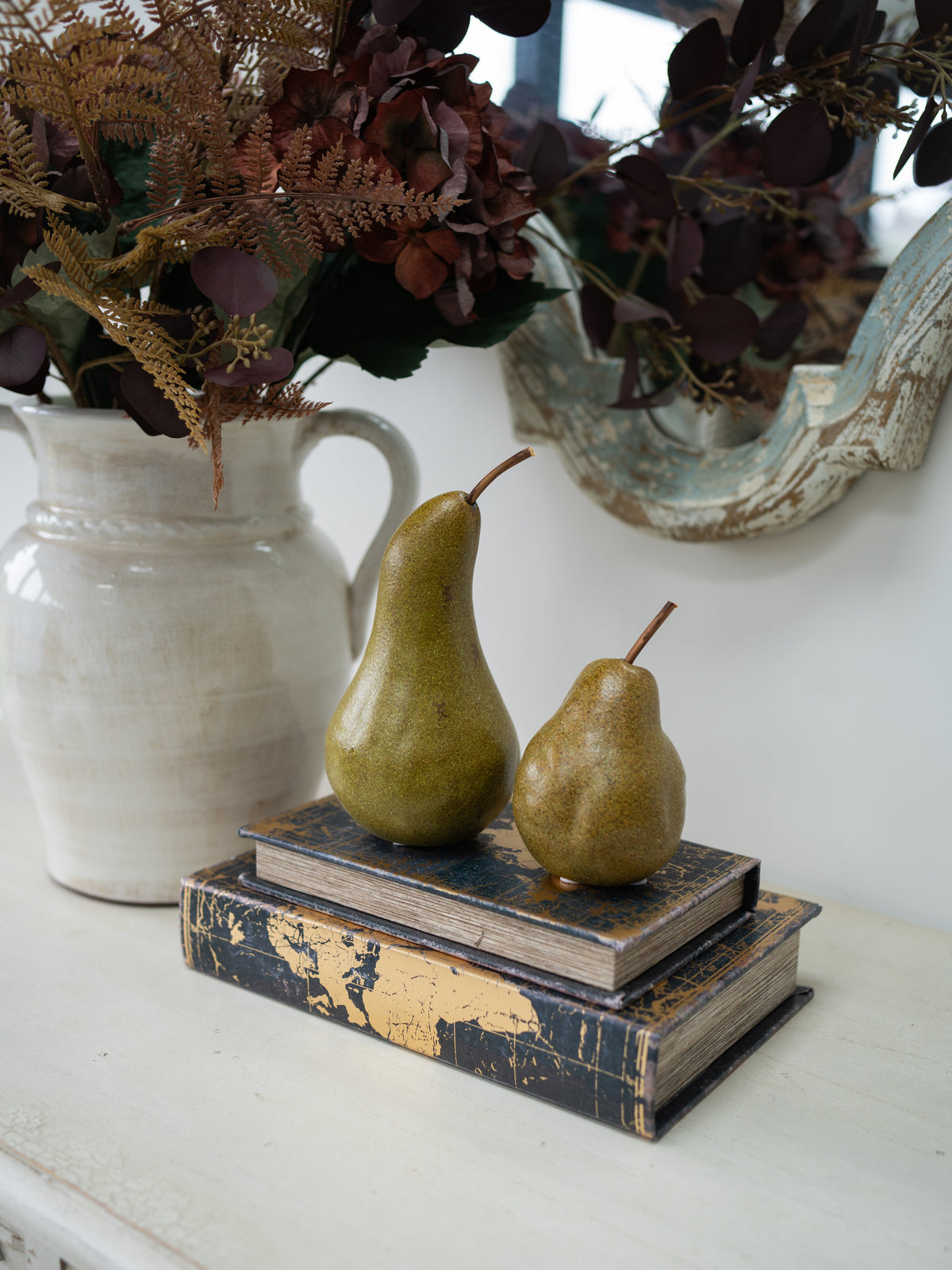 Two decorative pears placed on top of stacked vintage-style books on a white tabletop, with a large cream pitcher and dried flowers in the background.