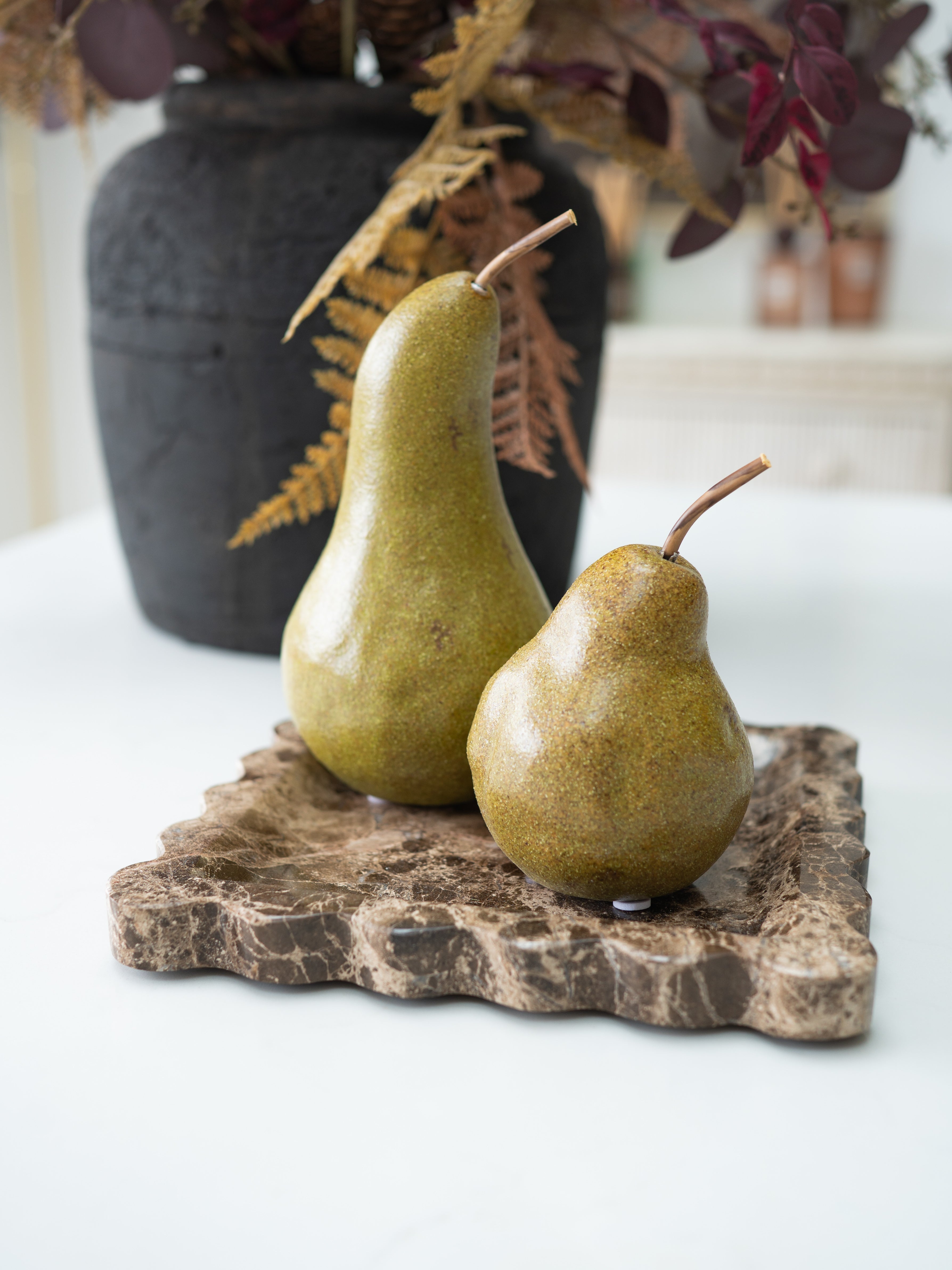 Two decorative pears with a glossy finish resting on a rustic stone tray, with a dark textured vase and dried foliage in the background.
