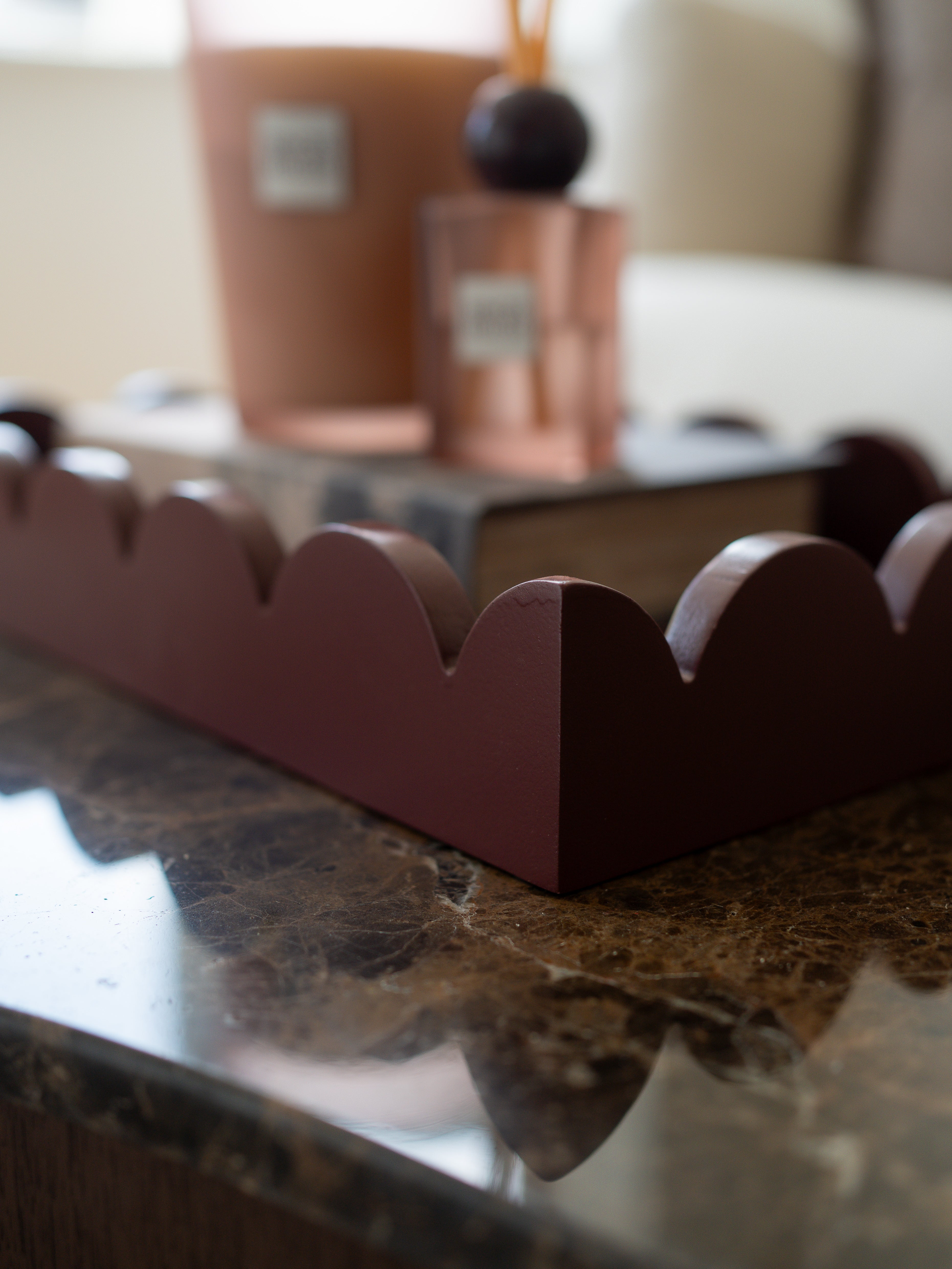 A scalloped-edge wooden tray in a deep reddish-brown finish, styled on a polished dark marble countertop with a blurred background showing home decor items.