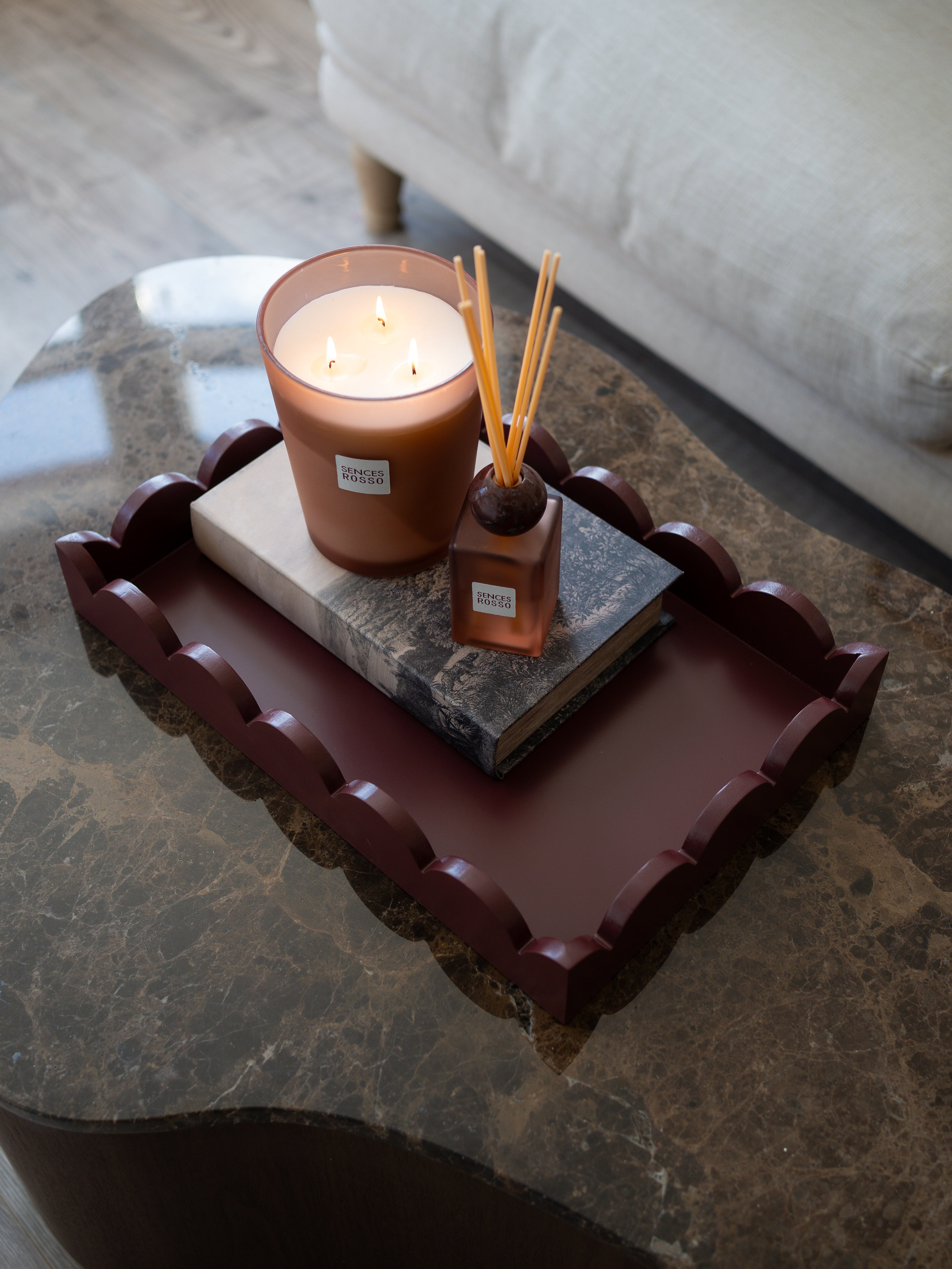 A styled home interior scene showing a dark reddish-brown scalloped wooden tray holding a lit candle and a reed diffuser on top of stacked books, placed on a marble coffee table with a sofa in the background.