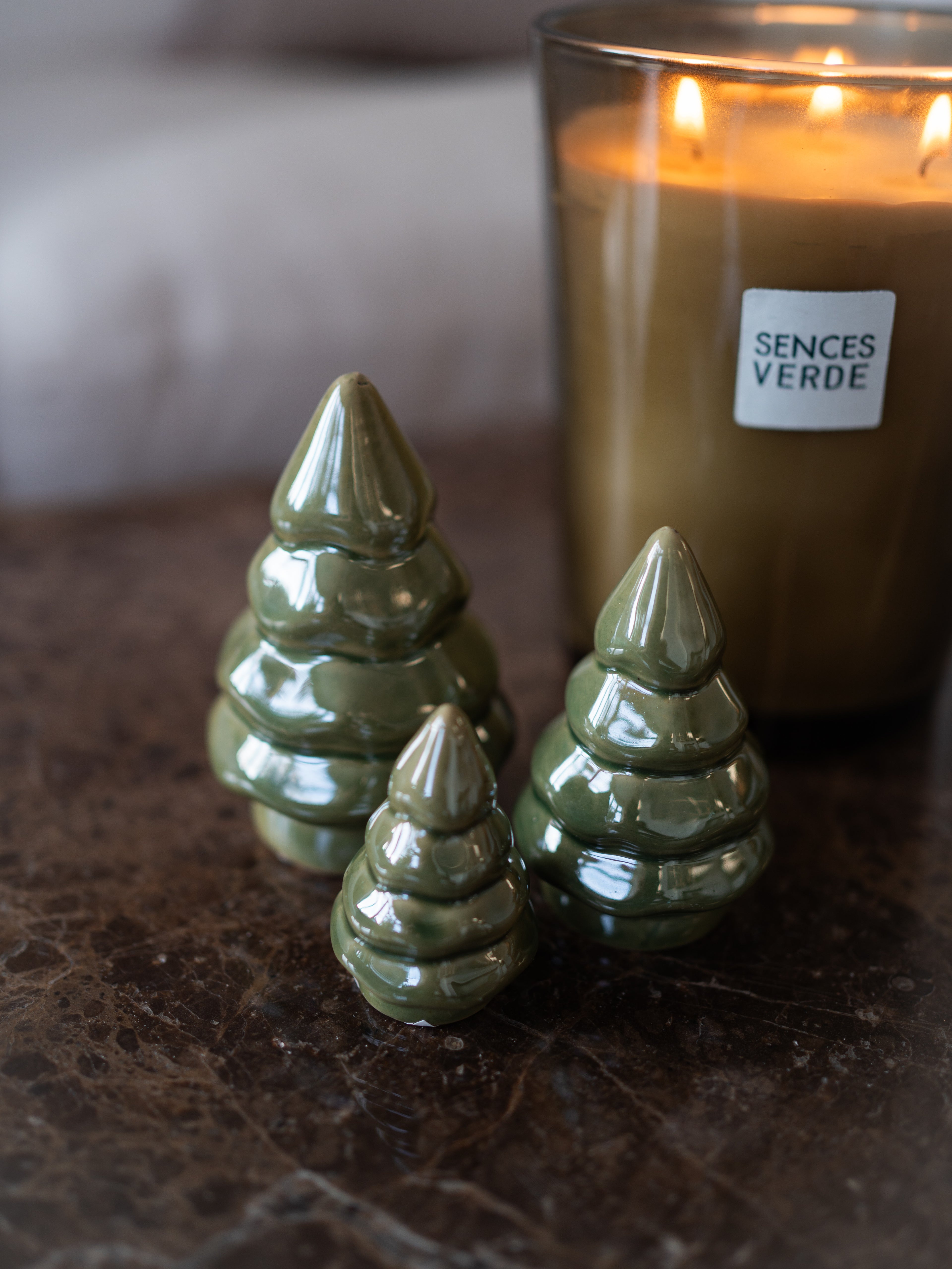 Three glossy pearl green ceramic Christmas tree ornaments arranged on a dark marble surface in front of a lit candle labeled &