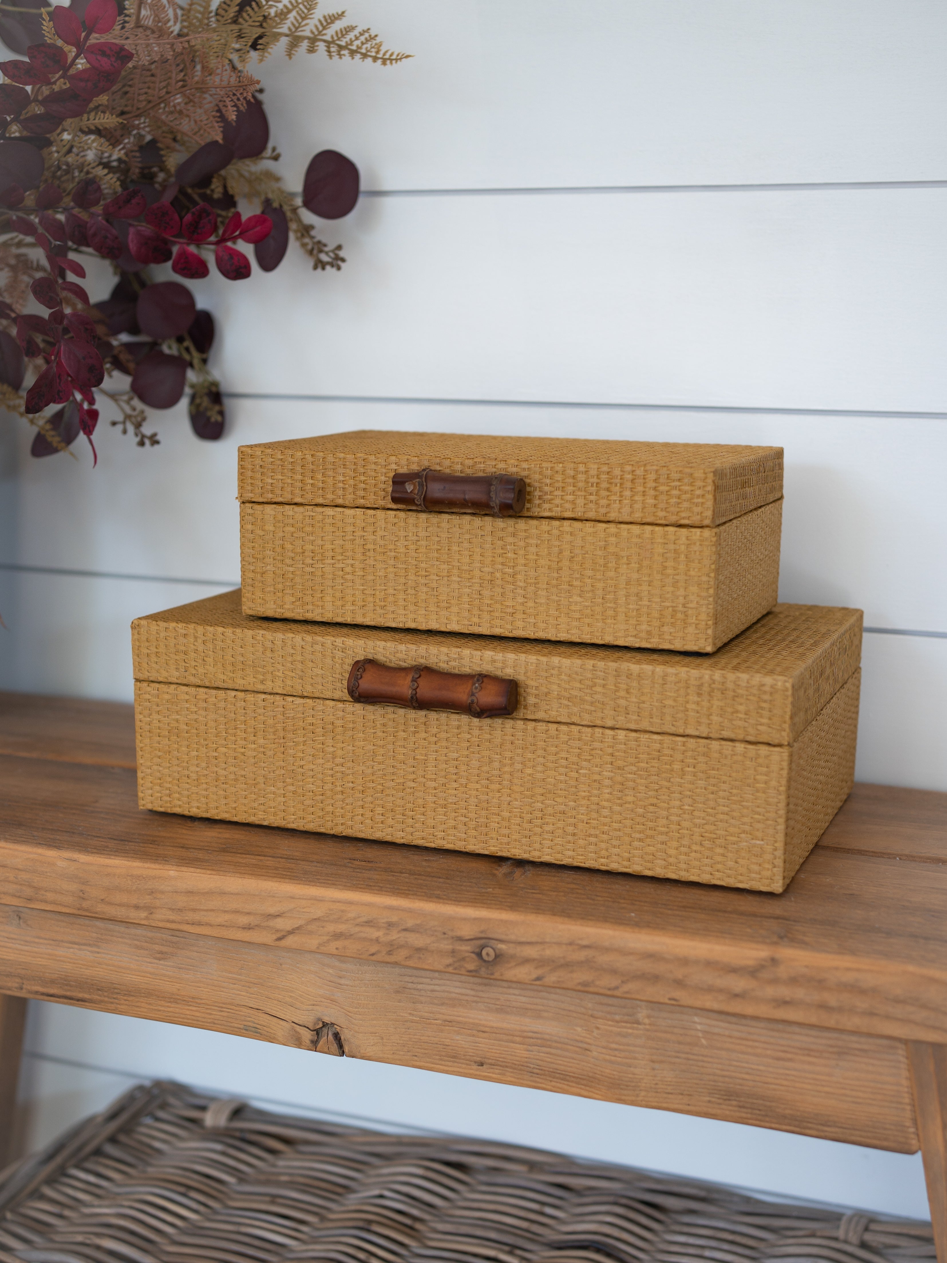 Two stacked woven storage boxes in a warm tan color with dark brown wooden bamboo handles, displayed on a rustic wooden bench against a white shiplap wall.