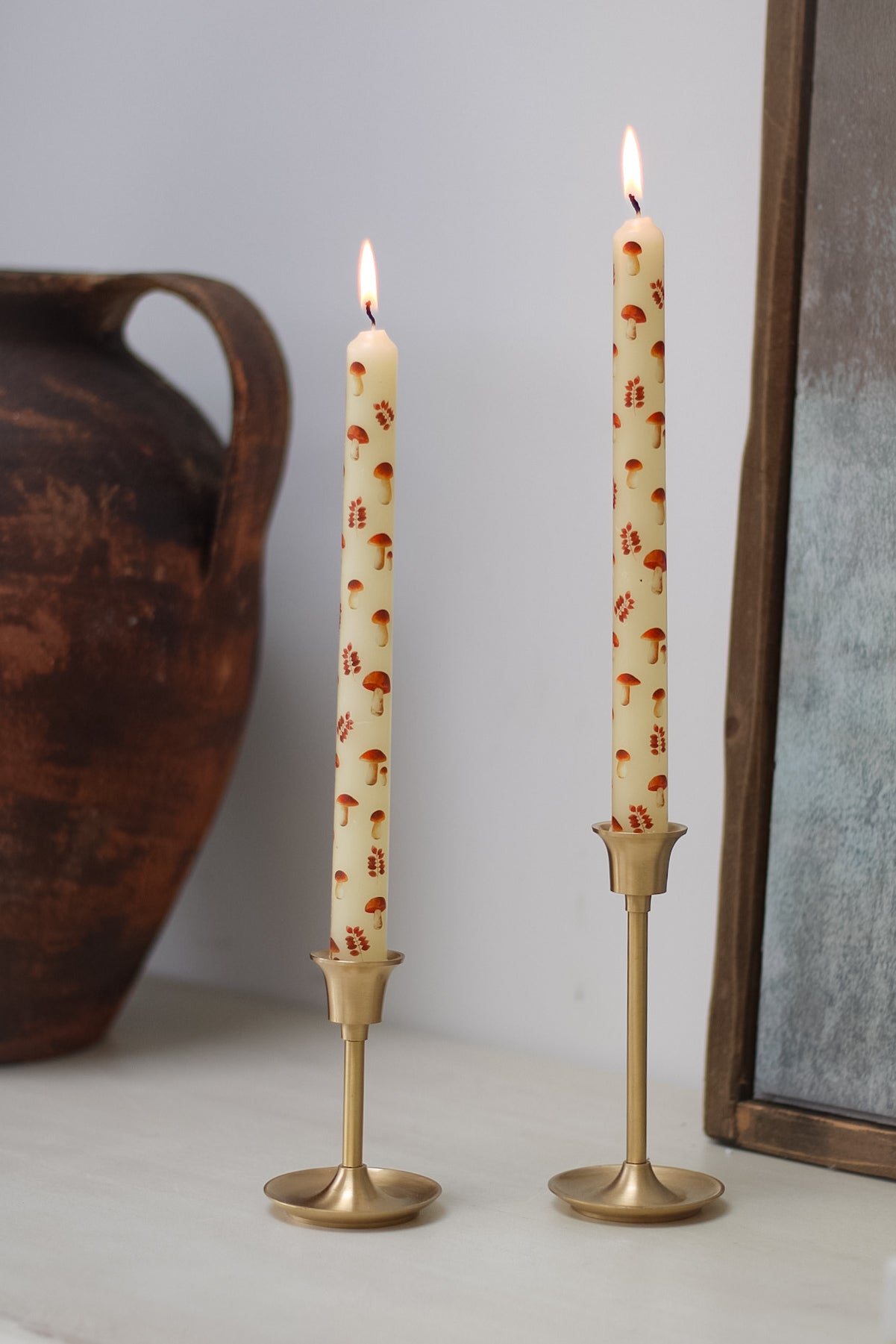Two lit toadstools and leaves candles in brass candlesticks on a neutral background