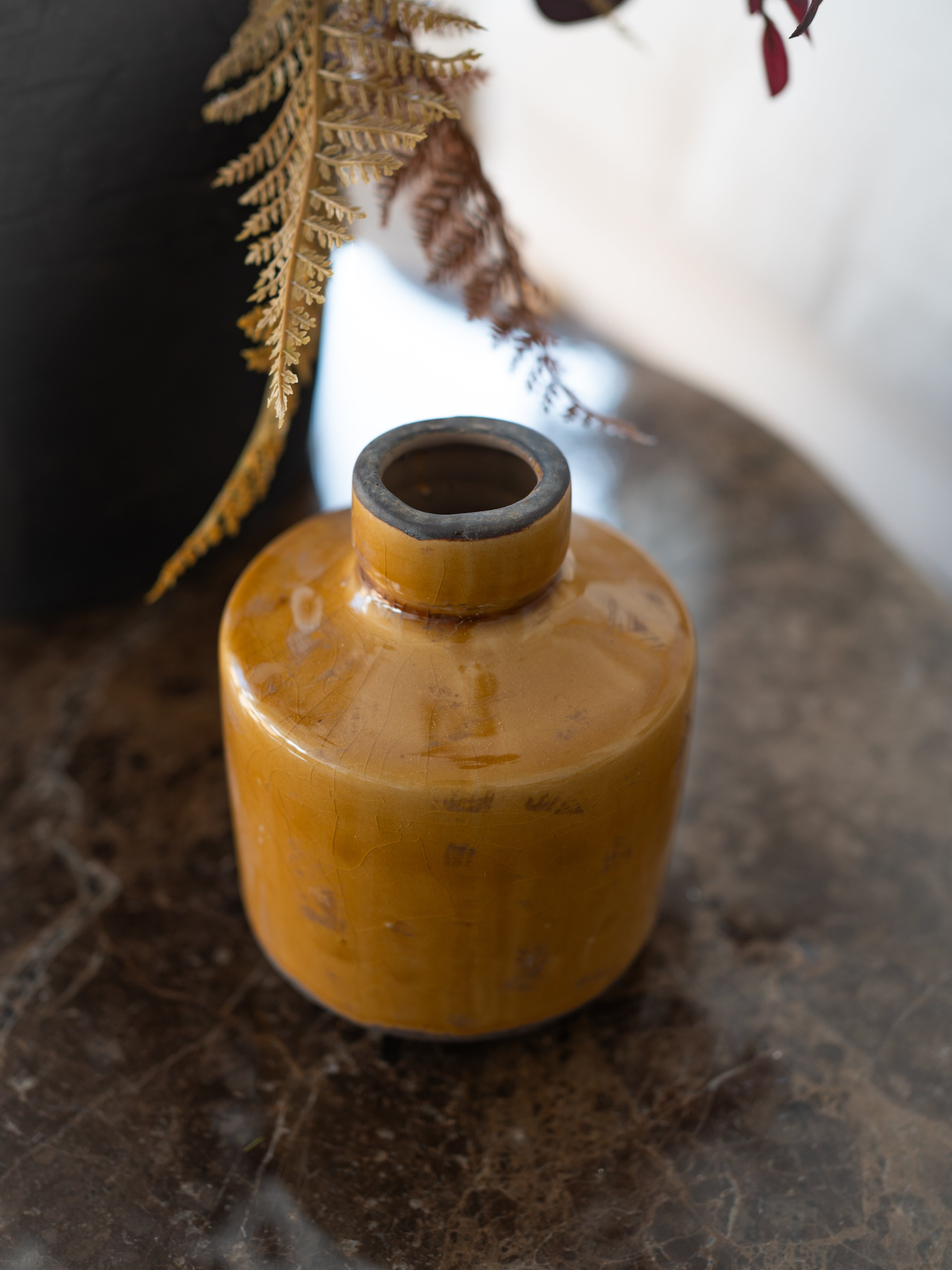 A small cylindrical ceramic vase with a glossy mustard-yellow glaze and a dark rim, styled on a dark stone surface with dried ferns in the background.