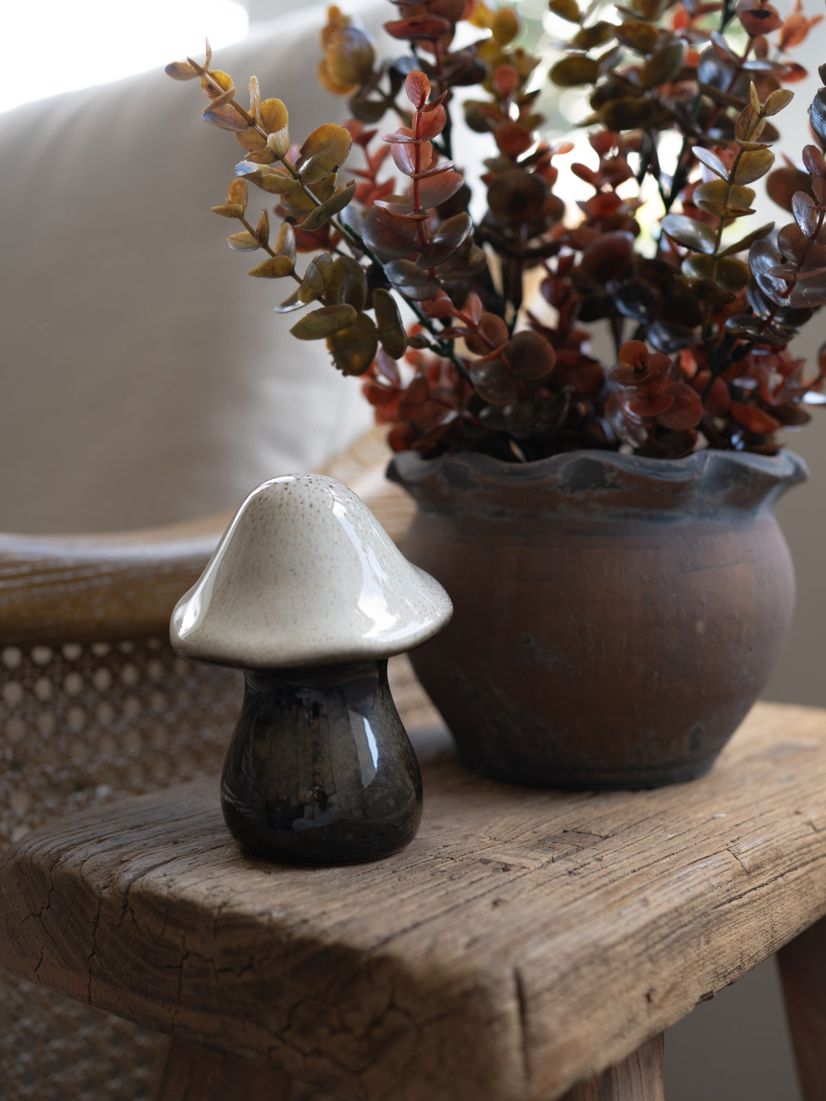 A small ceramic mushroom-shaped decor piece with a glossy white cap and dark glazed base, displayed on a rustic wooden table next to a potted plant.