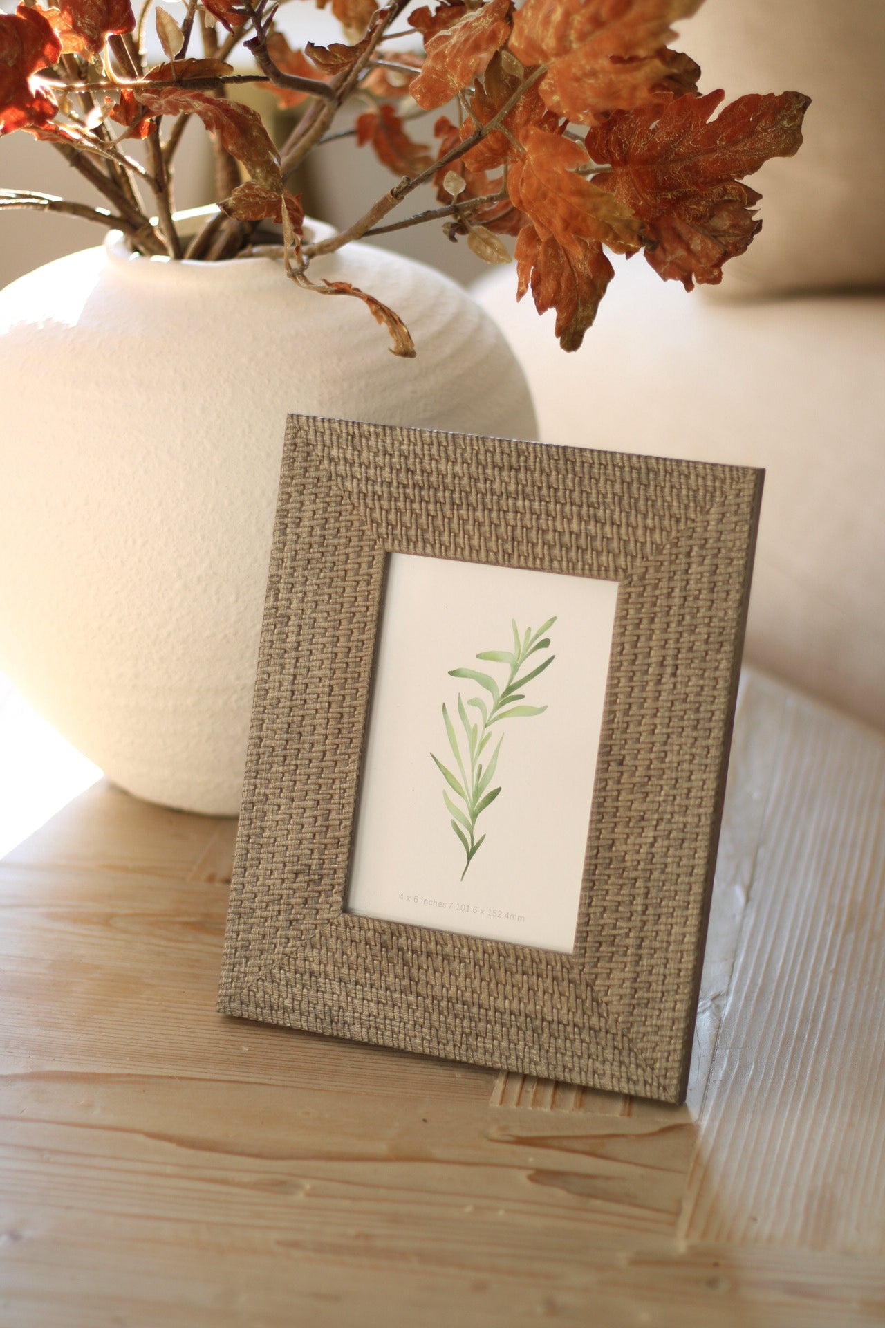 Soft grey woven photo frame with a plant on a wooden surface next to a vase with autumn leaves.