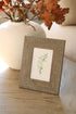 Soft grey woven photo frame with a plant on a wooden surface next to a vase with autumn leaves.