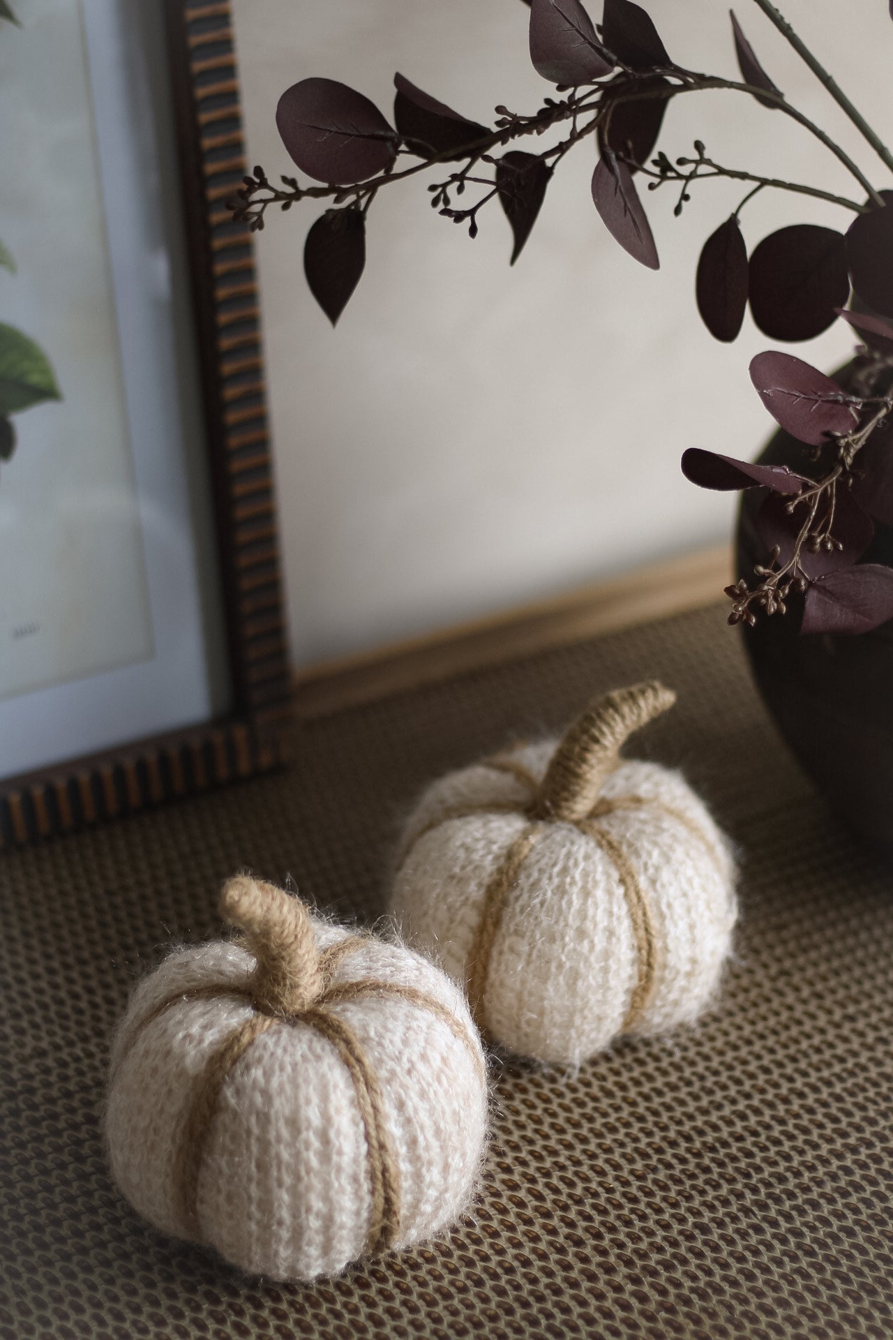 Two knitted pumpkins on a textured surface with a framed picture and plant in the background.