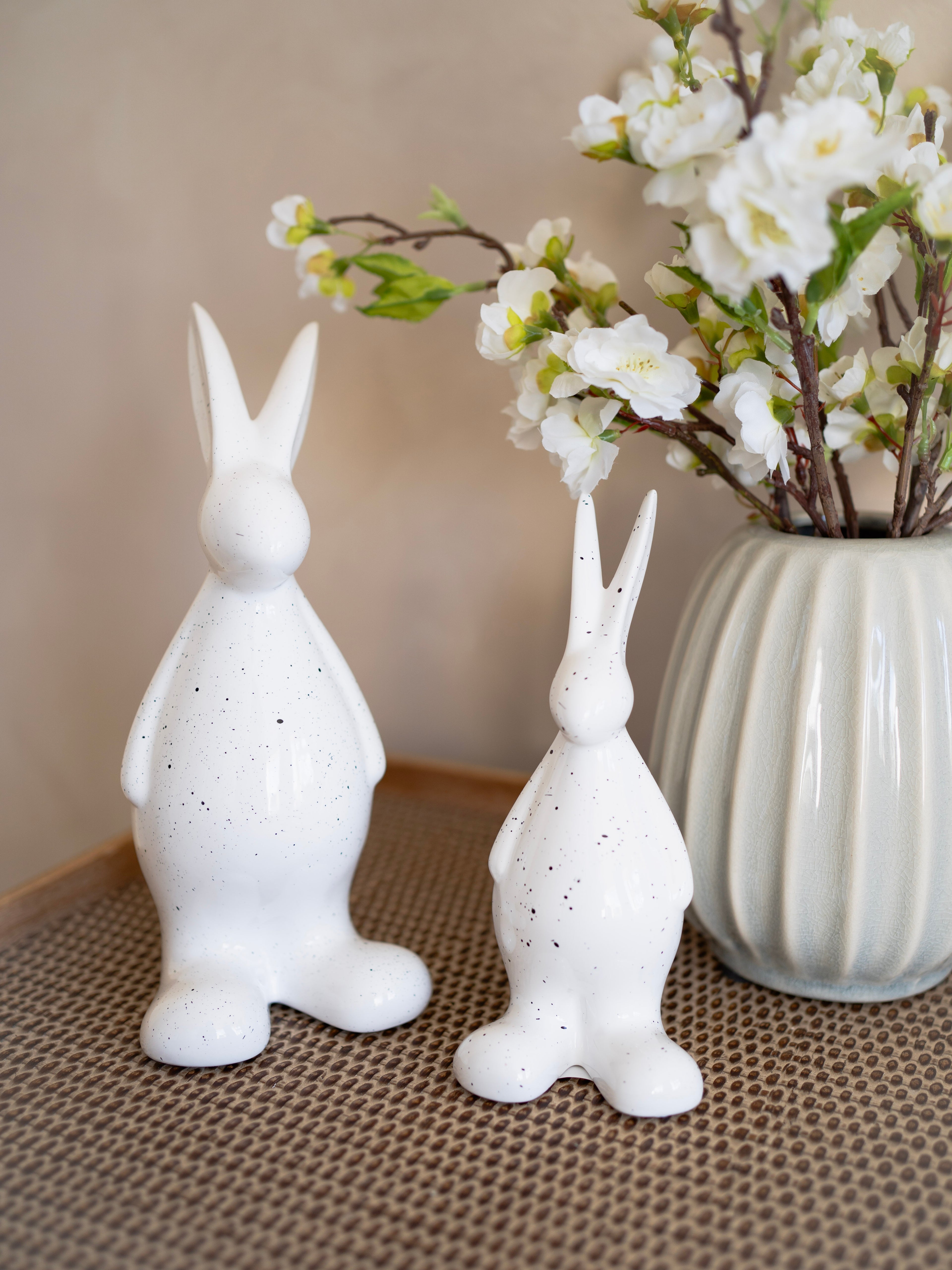 Two white ceramic rabbit sculptures on a textured surface with a vase of flowers in the background.