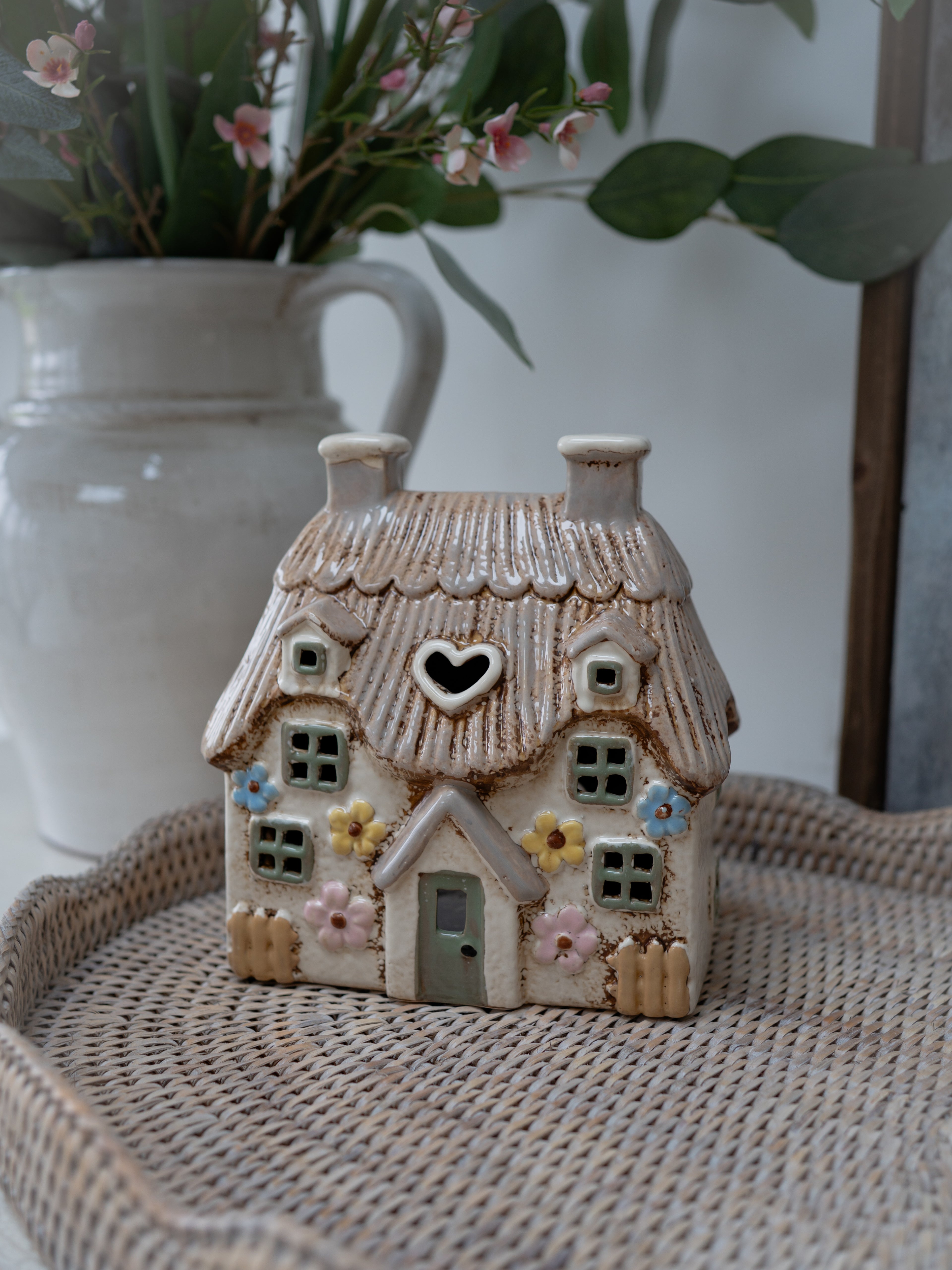 Small ceramic house with a heart window on a woven surface with a blurred vase and flowers in the background.