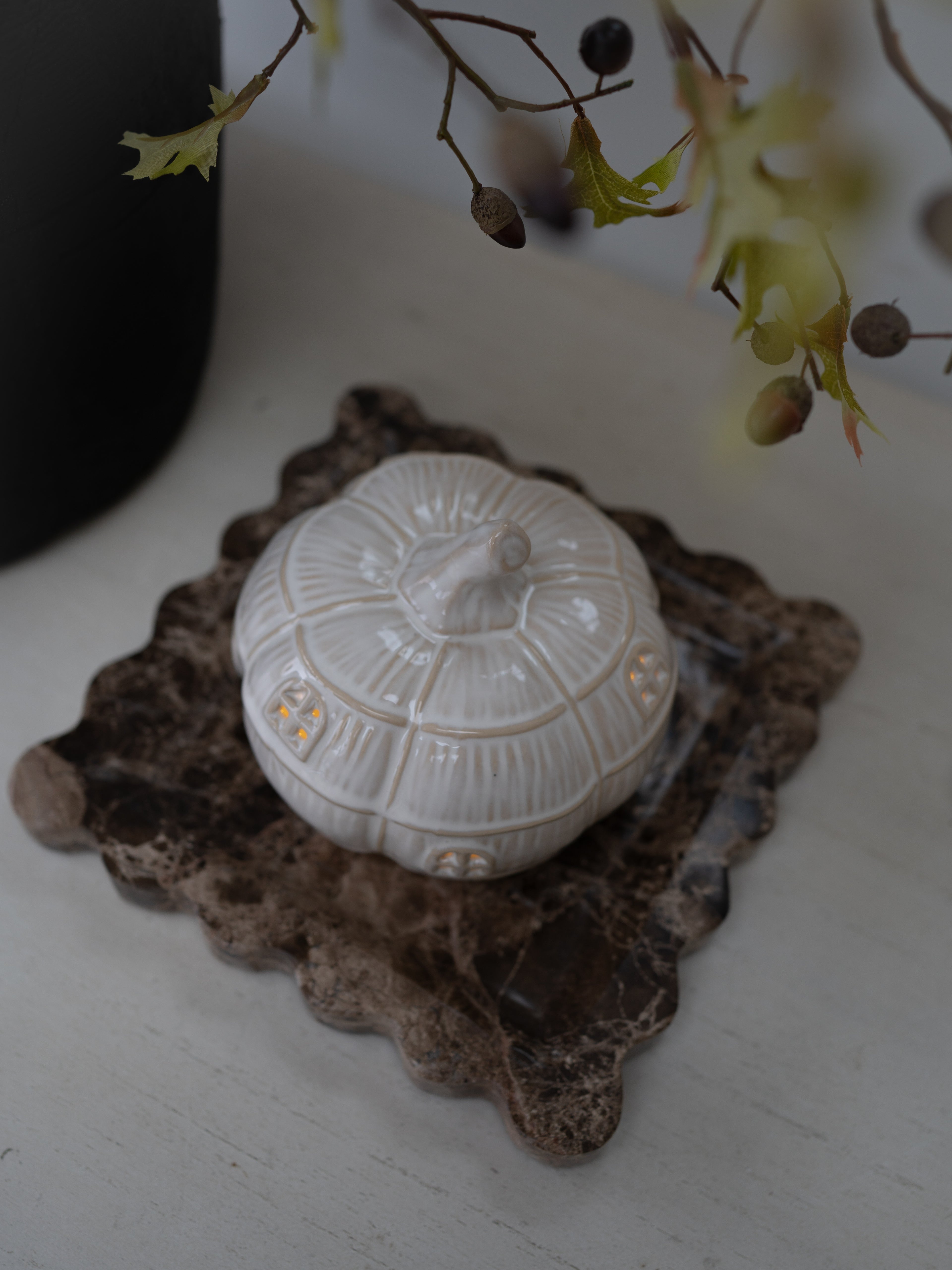 A small white ceramic pumpkin-shaped ceramic ornament with subtle gold accents, displayed on a rustic brown stone coaster against a light gray surface.