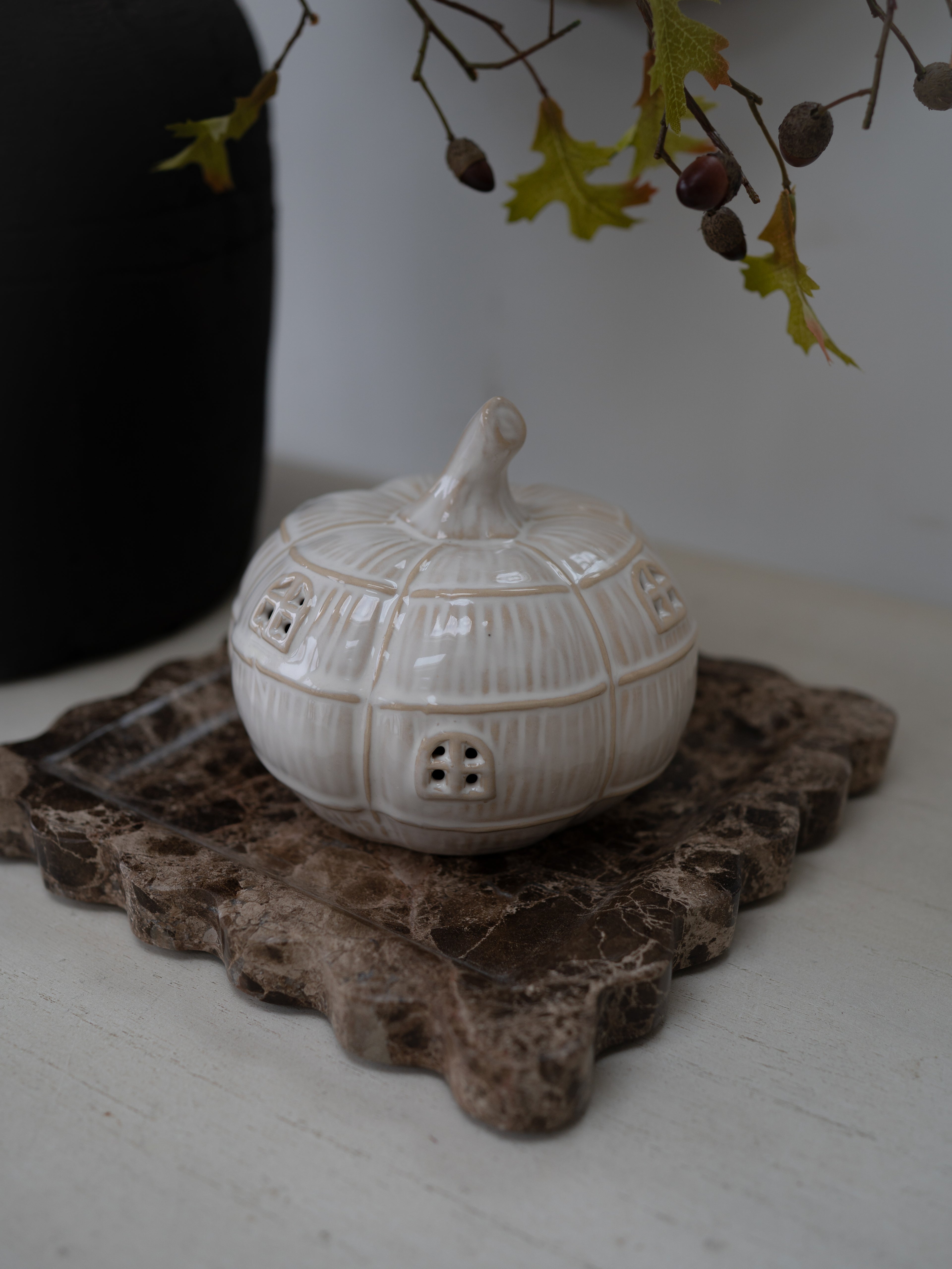 A small cream-colored ceramic house-shaped container with a lid, displayed on a rustic brown stone slab with a blurred black vase and autumn leaves in the background.