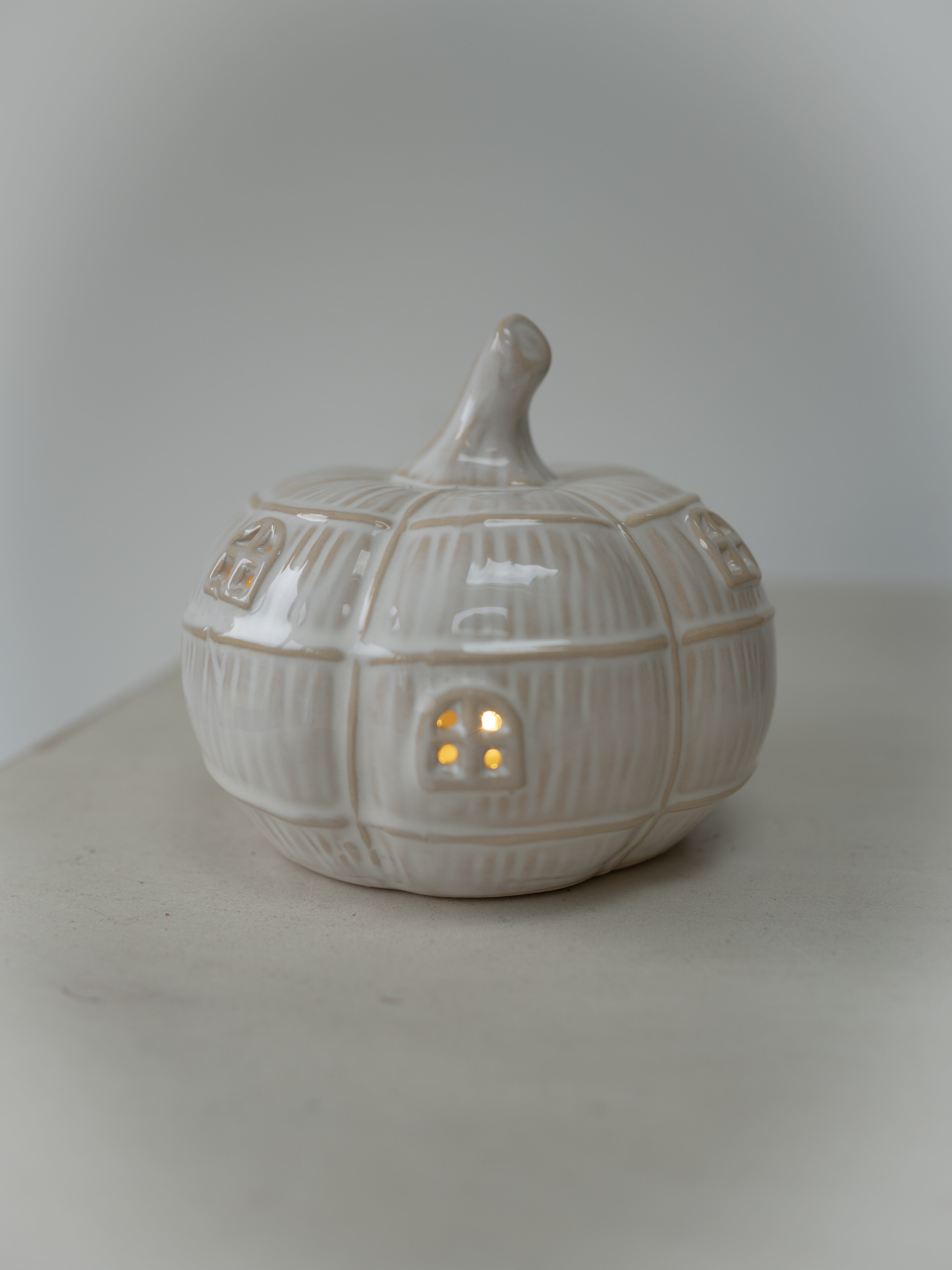A small ceramic pumpkin-shaped house with a lit warm yellow window, shown on a neutral light gray background.