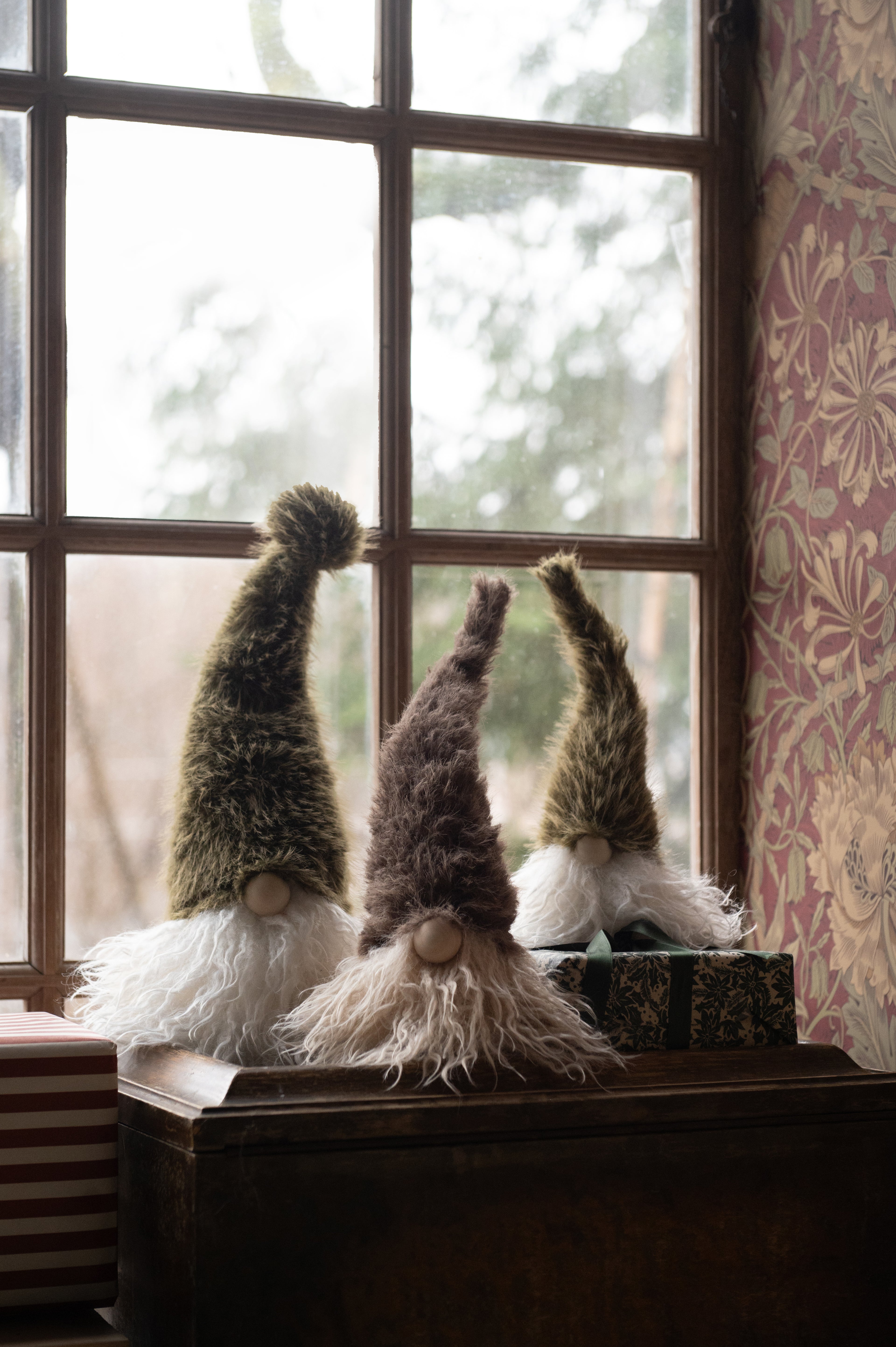 Three fuzzy Christmas gonk decorations with tall pointed hats and white beards sit on a dark wooden chest in front of a window. The gnomes have textured olive green, brown, and tan hats with white and cream beards, and one is posed behind a wrapped gift box.