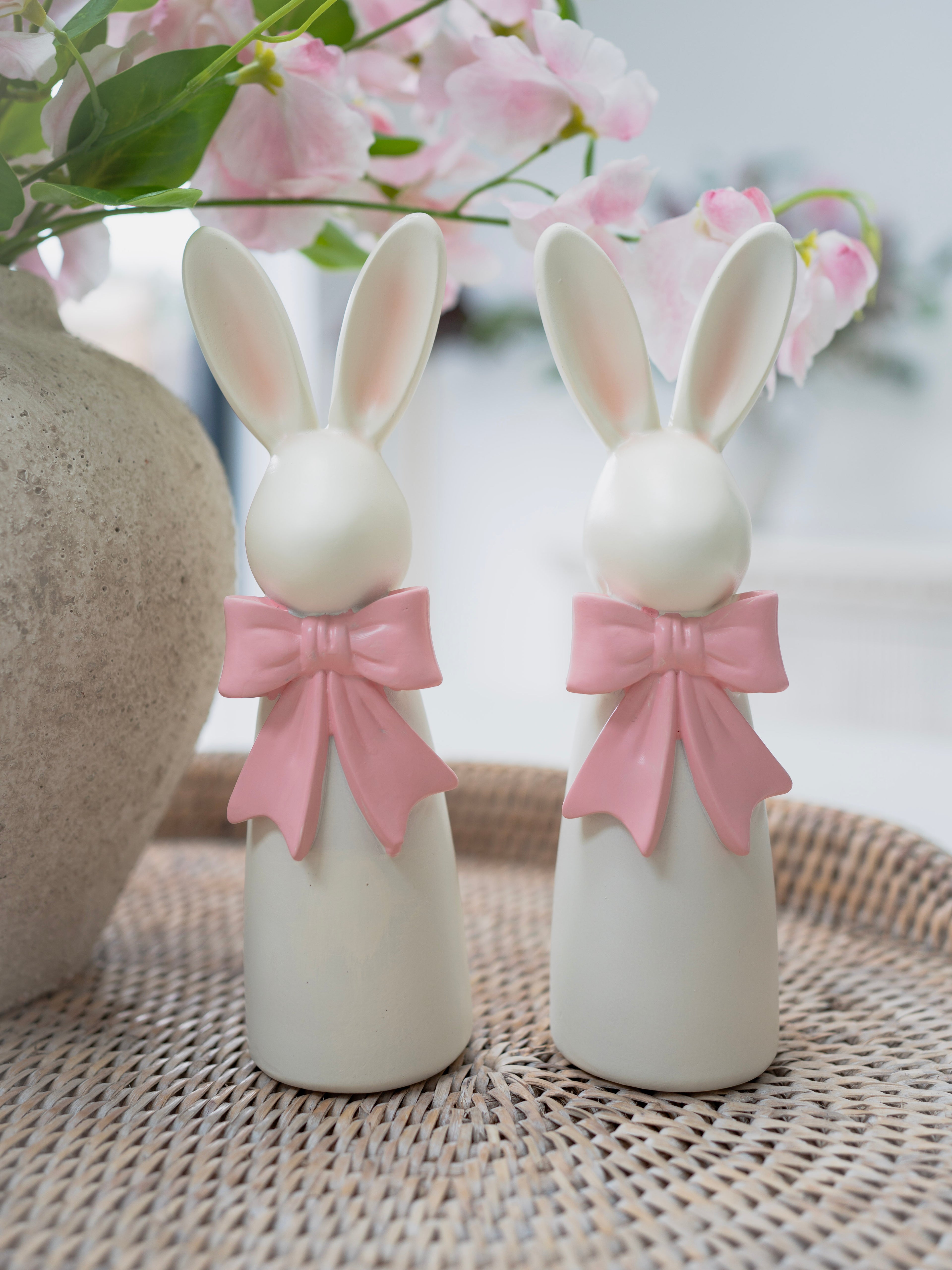 Two white rabbit figurines with pink bows on a woven surface with flowers in the background.