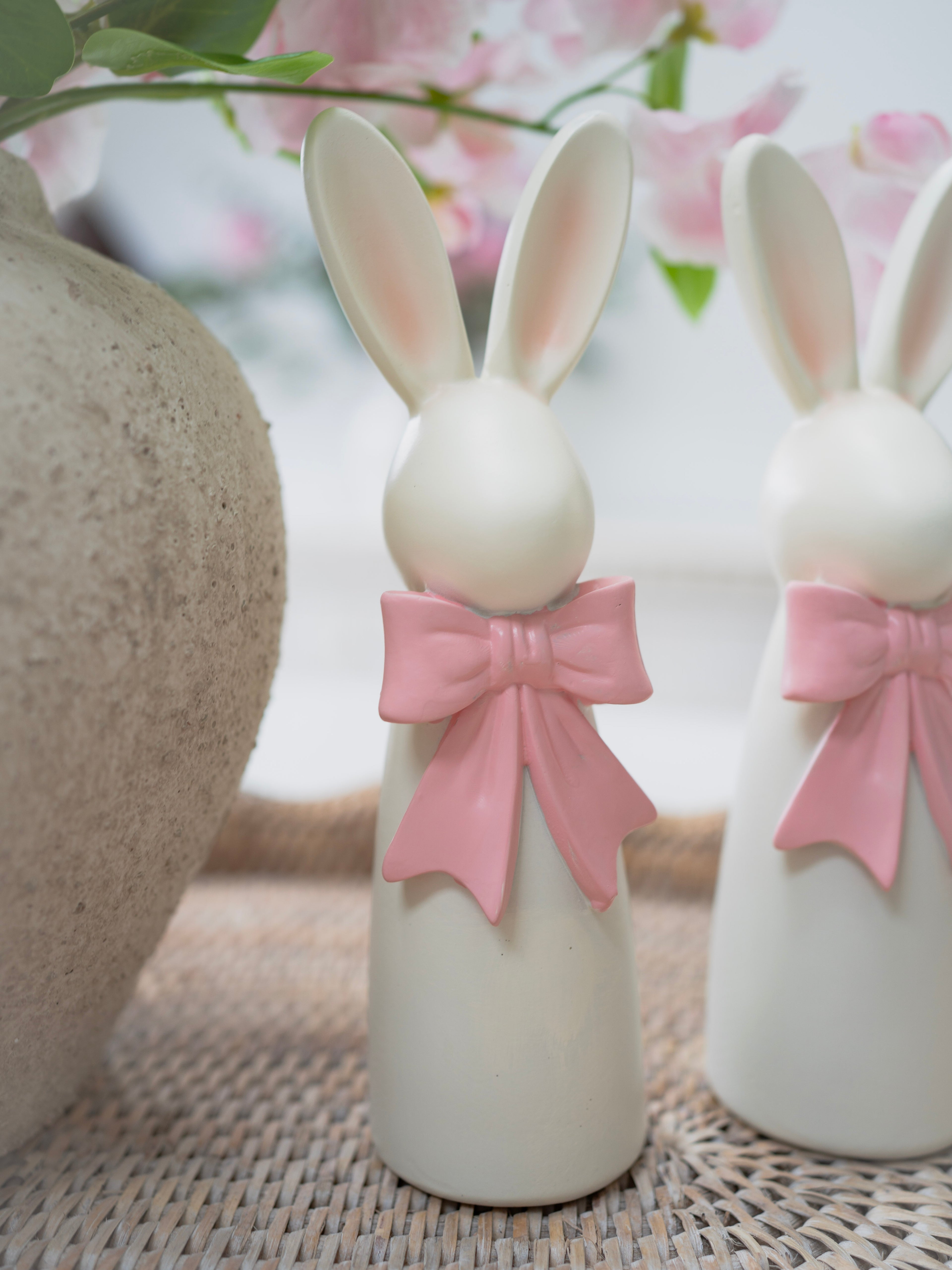 Two resin bunny figurines with pink bows on a textured surface with blurred floral background
