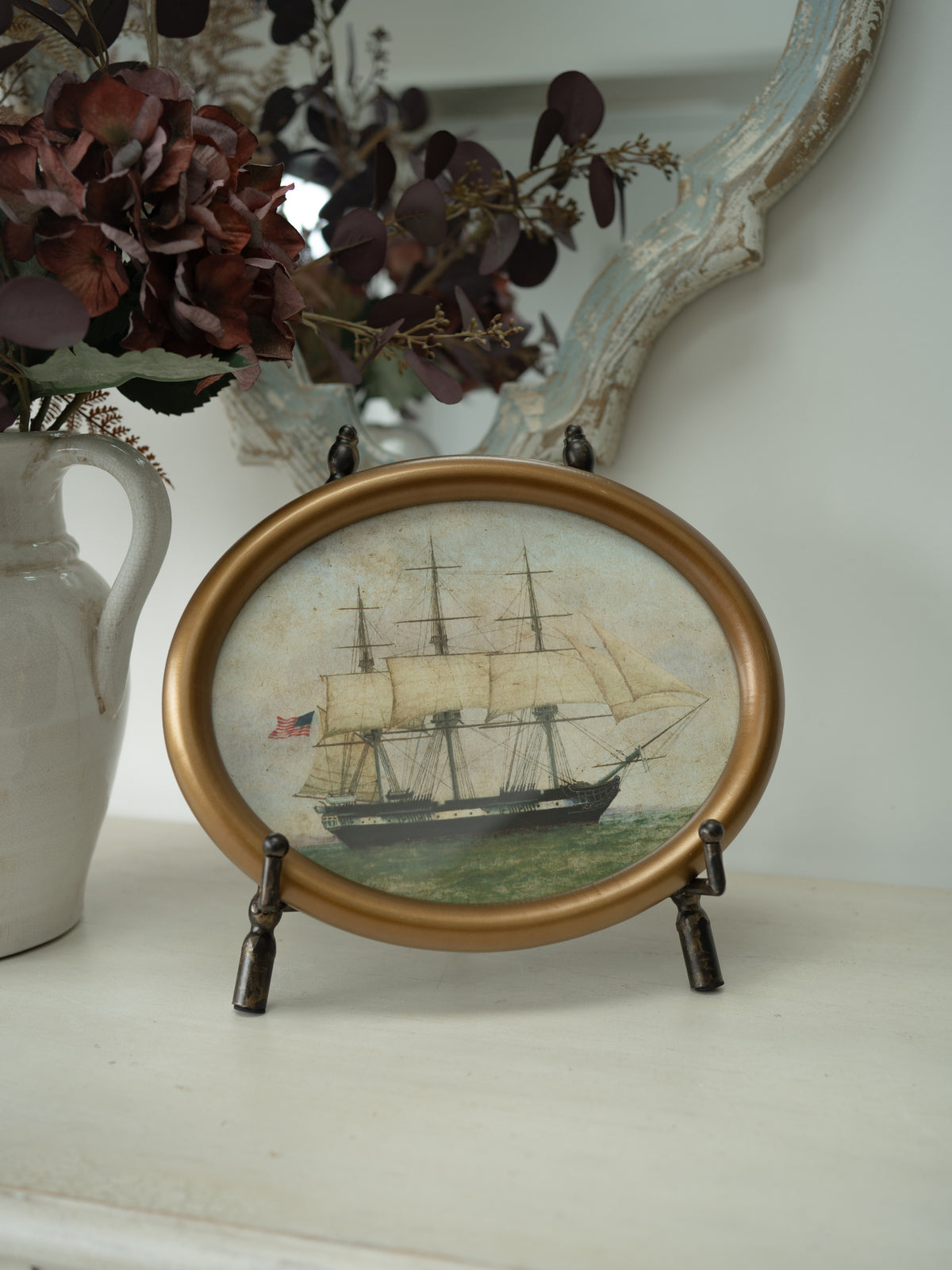 An oval framed miniature painting of a historic sailing ship on the water, displayed on a small dark metal stand on a white shelf next to a ceramic vase with dried flowers.