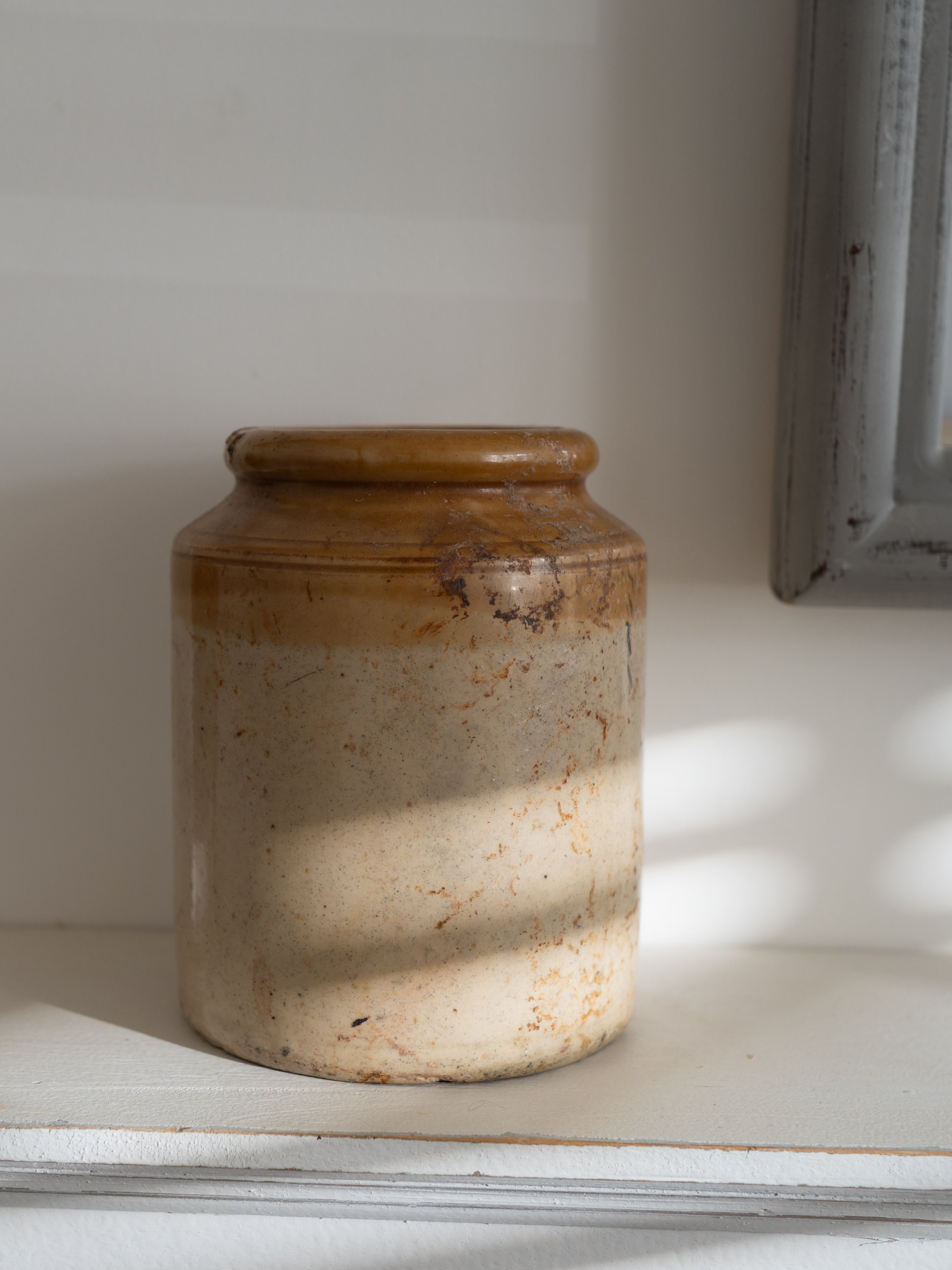 A rustic handmade ceramic vintage jar with a two-tone glaze, sitting on a white shelf in soft natural light.