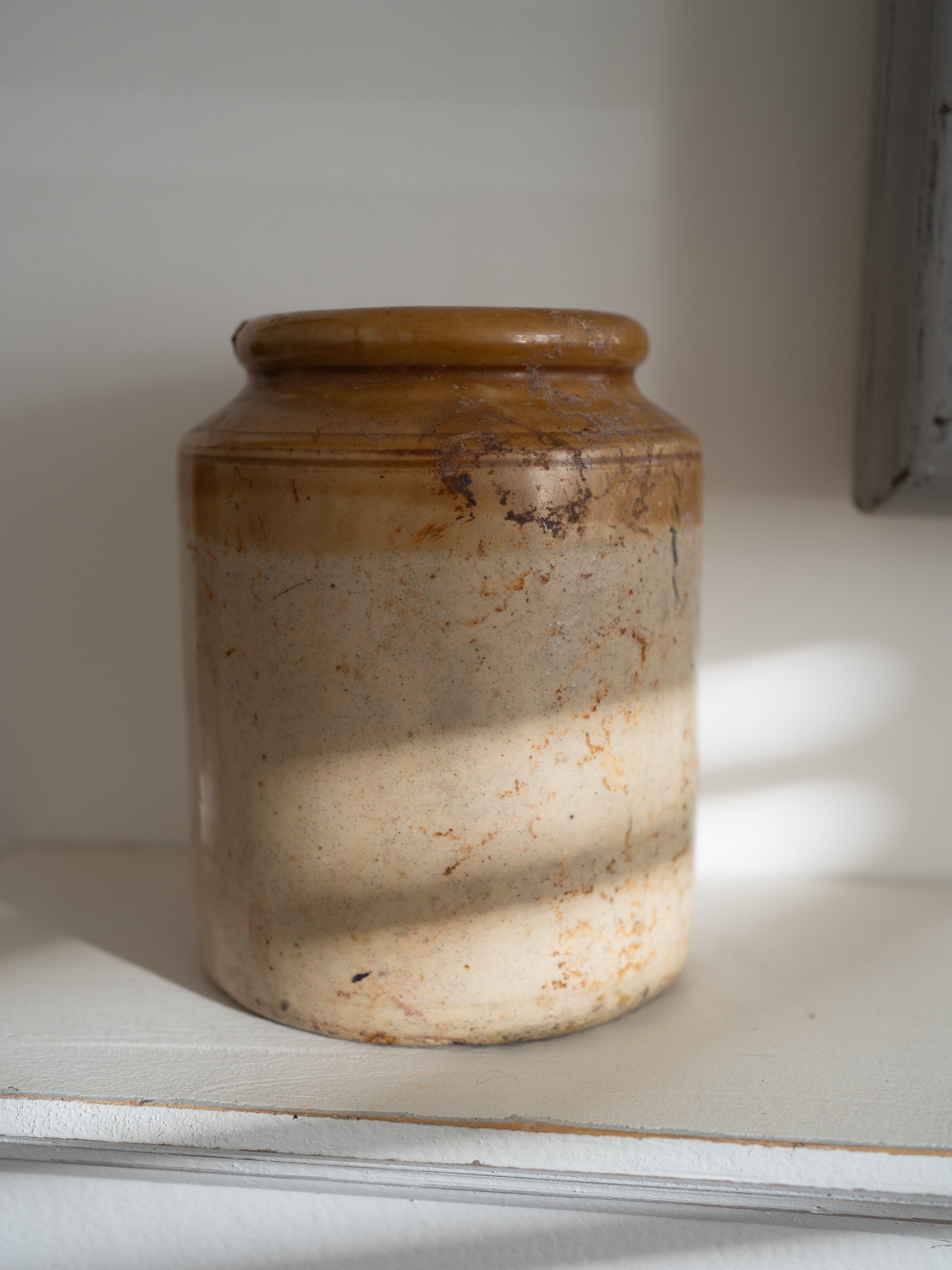 A rustic ceramic storage jar with a brown glazed top and a speckled beige lower body, sitting on a white shelf in soft natural light.