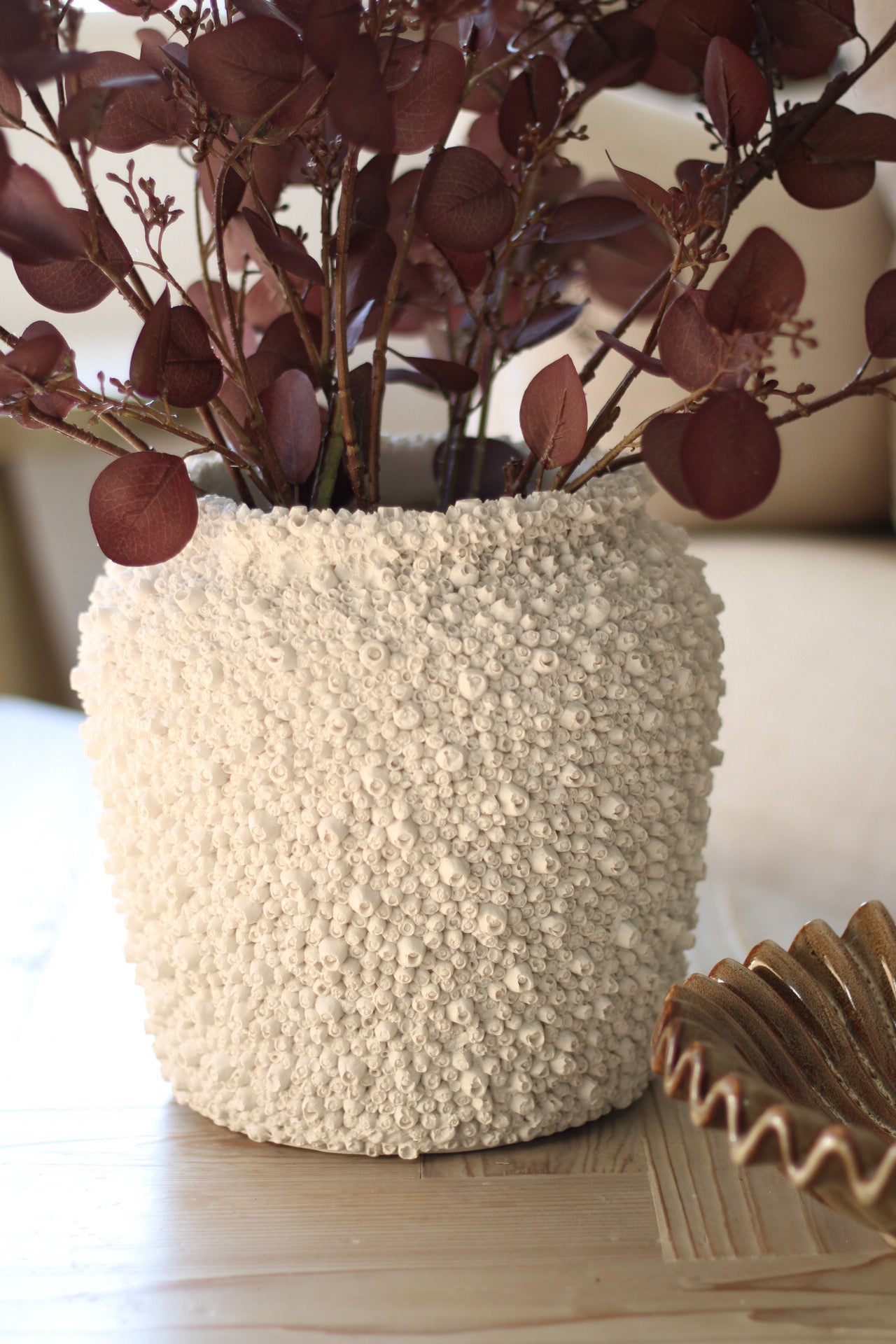 Decorative vase with textured barnacle surface holding purple foliage on a light wooden surface.
