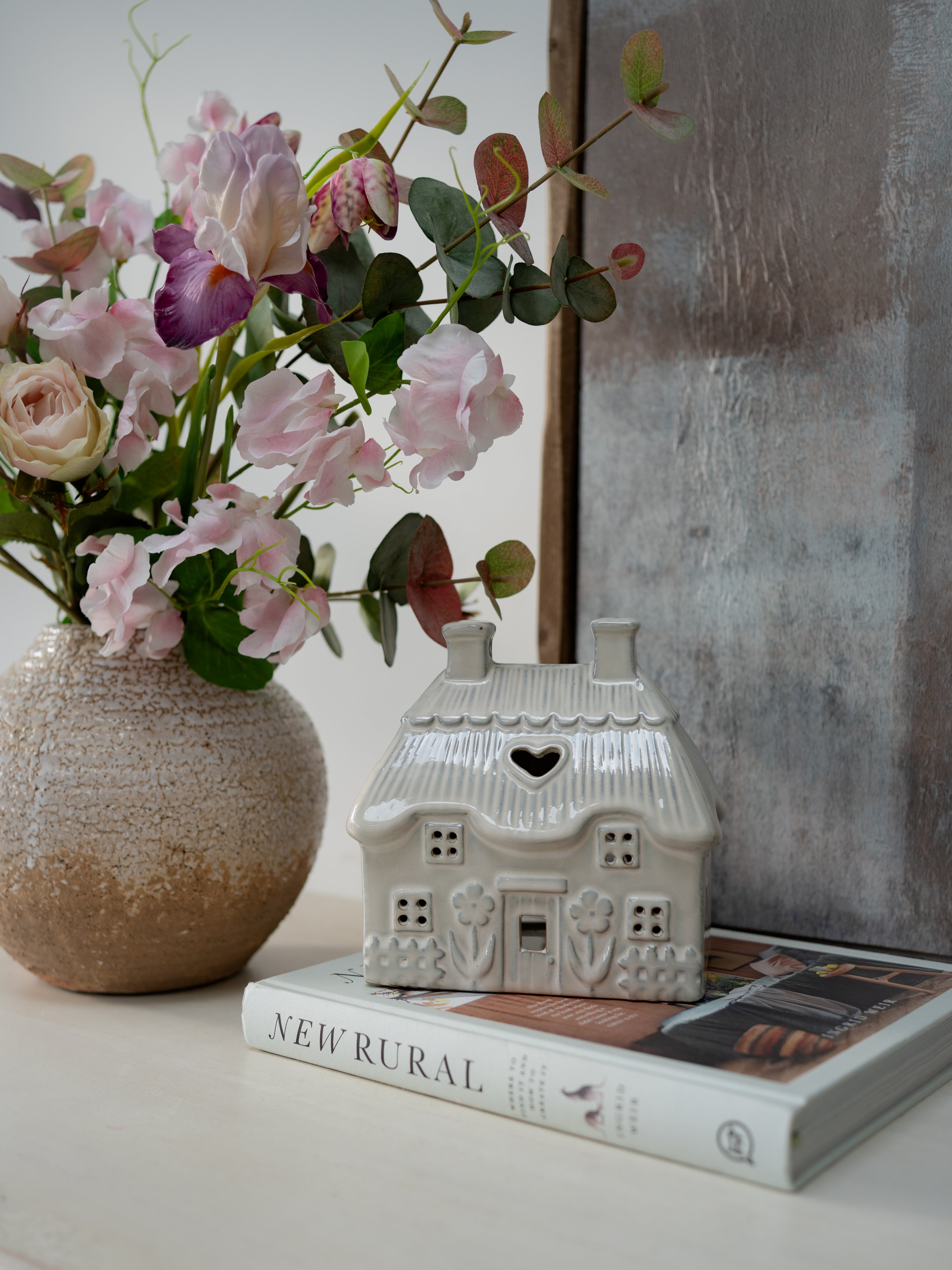 Decorative house-shaped object on a book with a vase of flowers and a mirror in the background