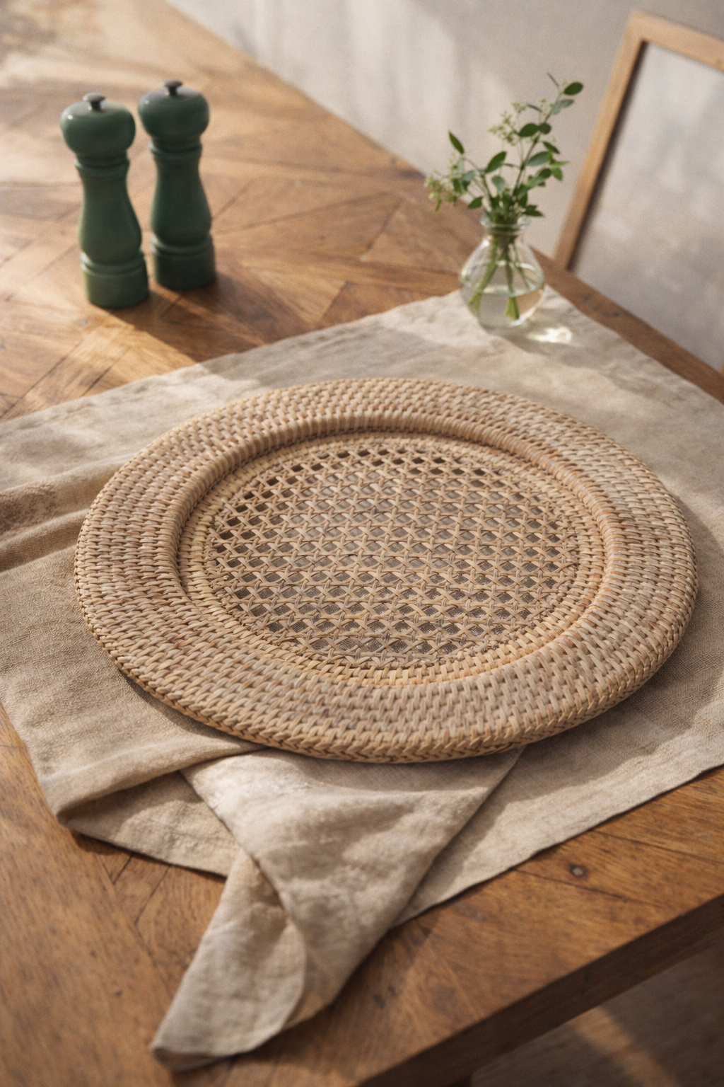 Round woven charger plate placemat on a wooden table with a salt and pepper shaker, and a vase with flowers in the background.