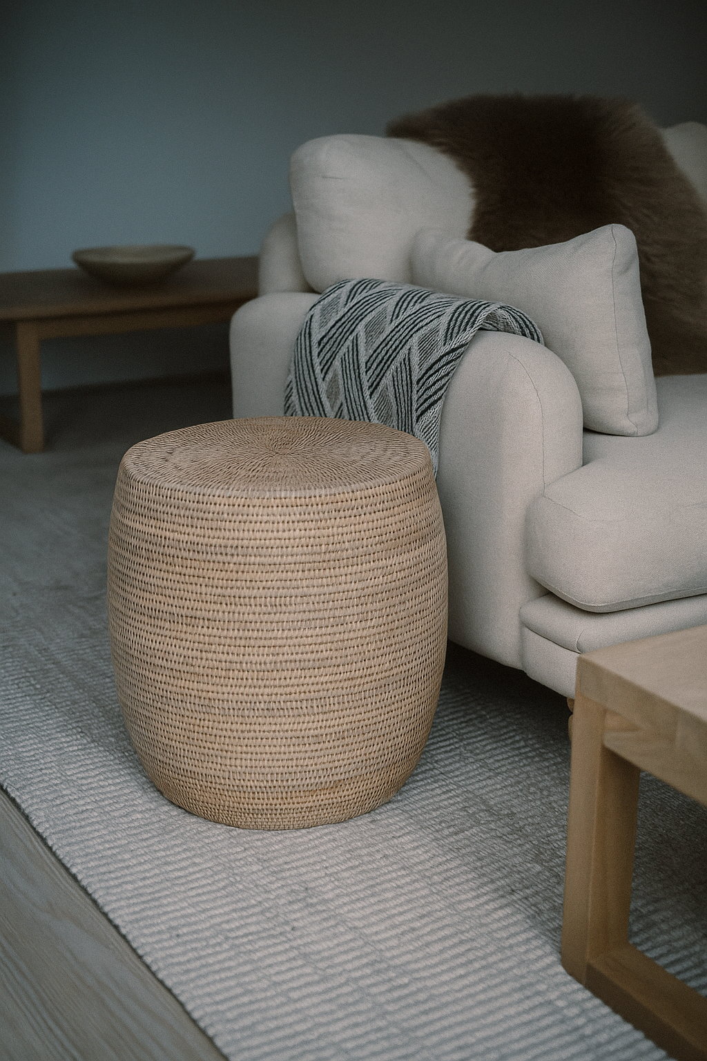 Woven rattan round drum stool ottoman in front of a beige sofa with decorative pillows in a living room setting.