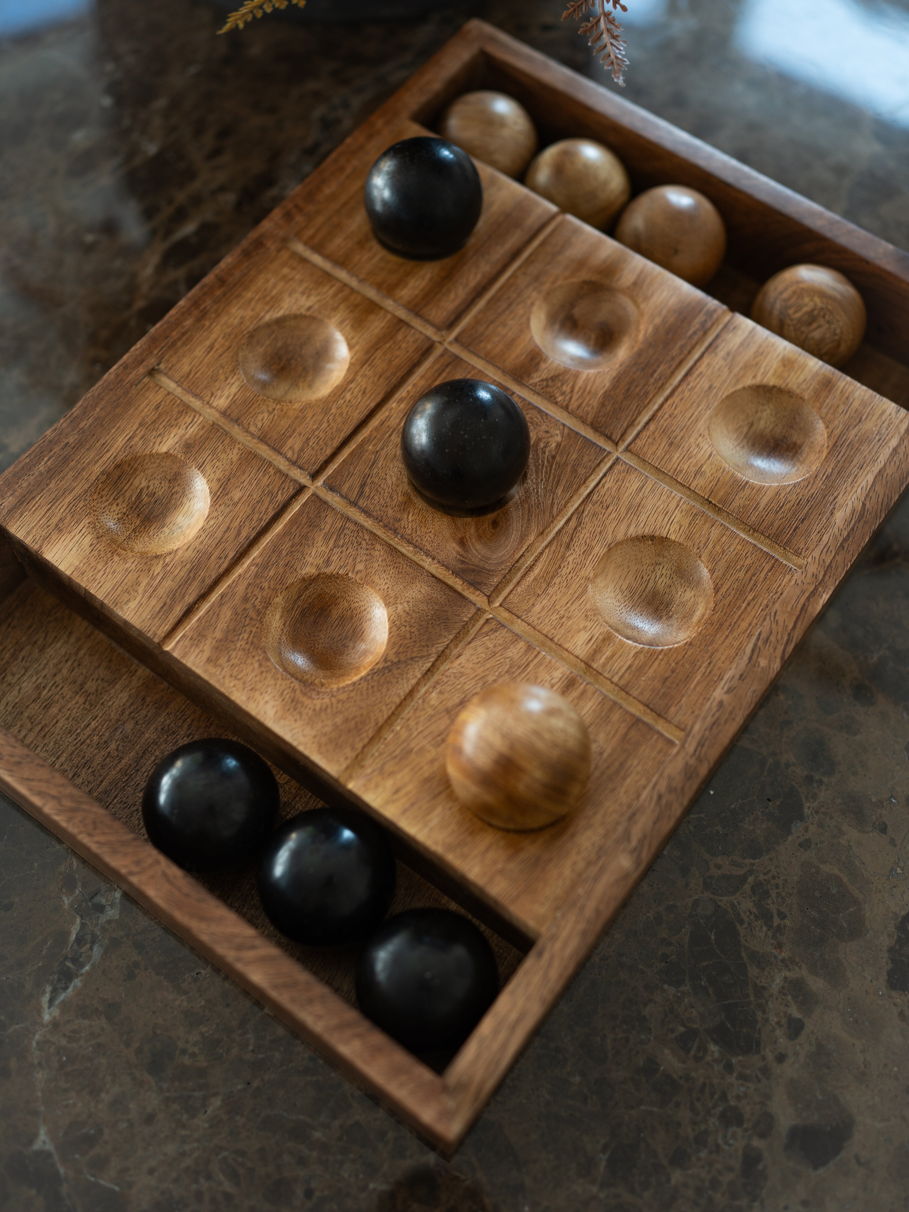 A wooden tic tac toe board game set with carved compartments and polished wooden balls, shown on a dark marble surface.