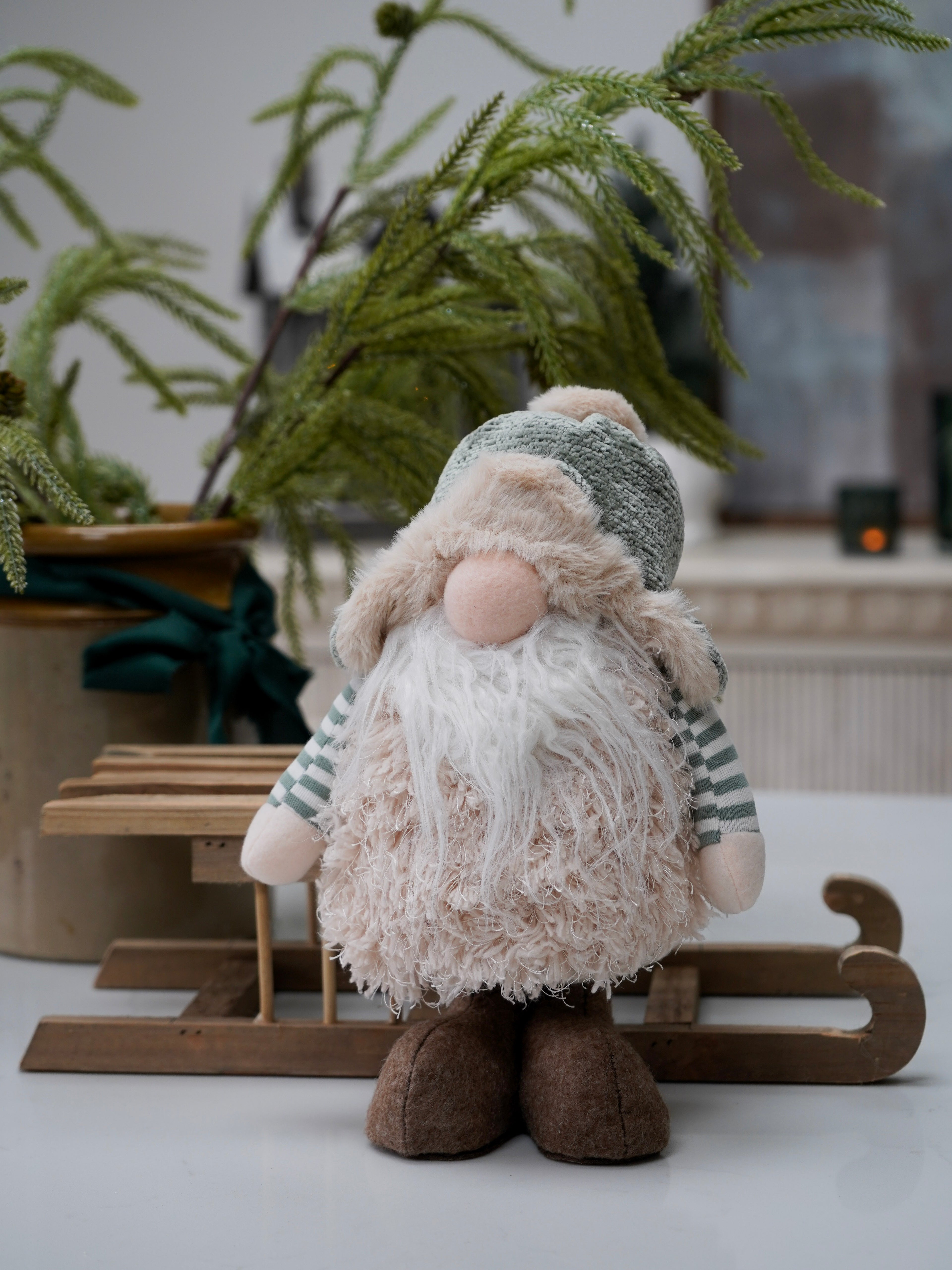 Decorative gnome gonk figure with a green hat and striped scarf, sitting on a wooden stand with plants in the background.