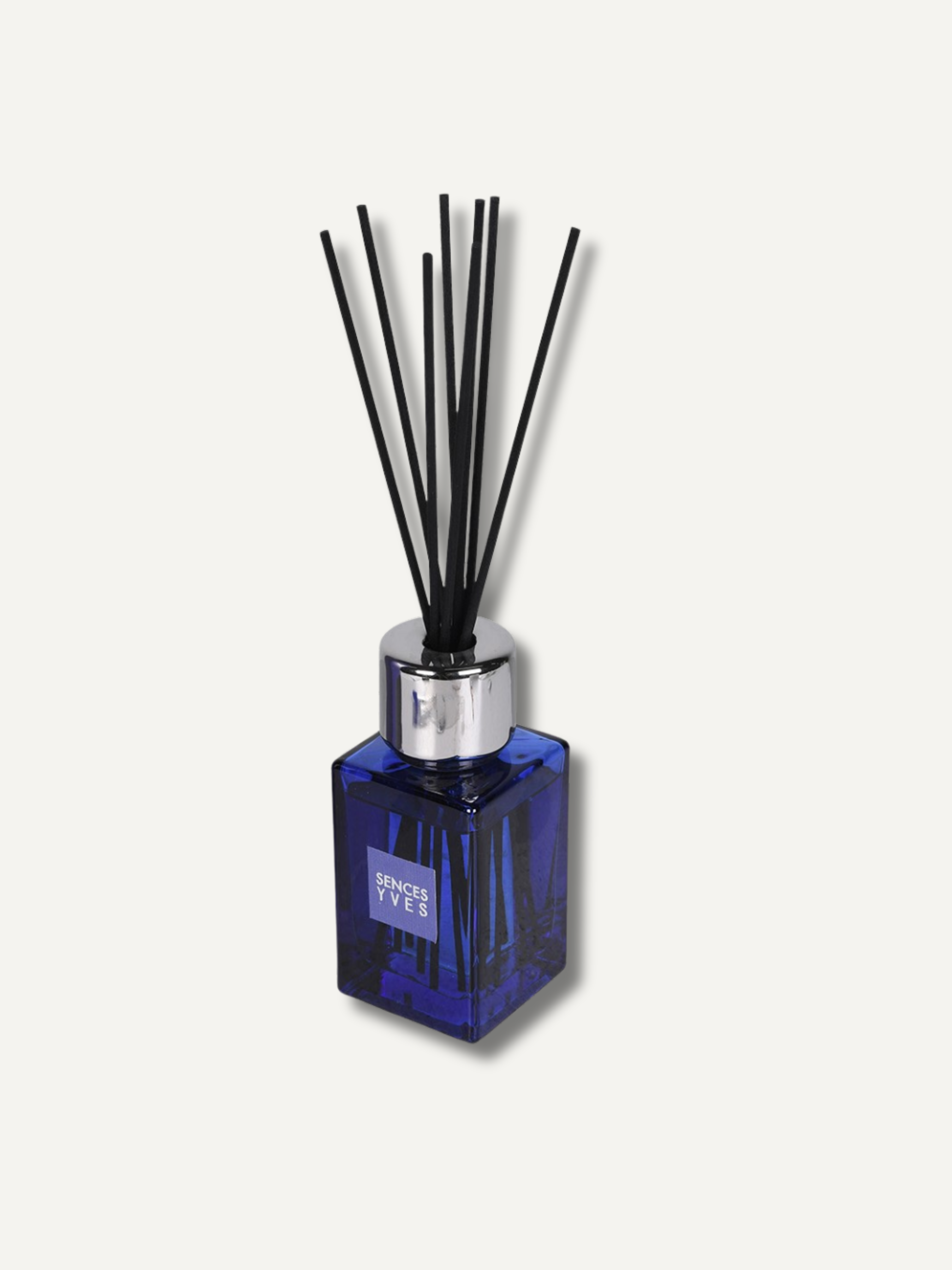 A reed diffuser bottle made of faceted cobalt blue glass with a shiny silver metal collar and multiple black reeds, shown on a light off-white background.