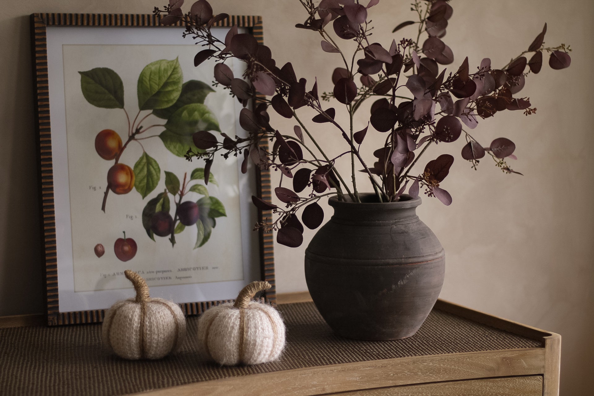 Decorative setup with a vase of purple faux eucalyptus, framed botanical print, and small pumpkins on a wooden surface.