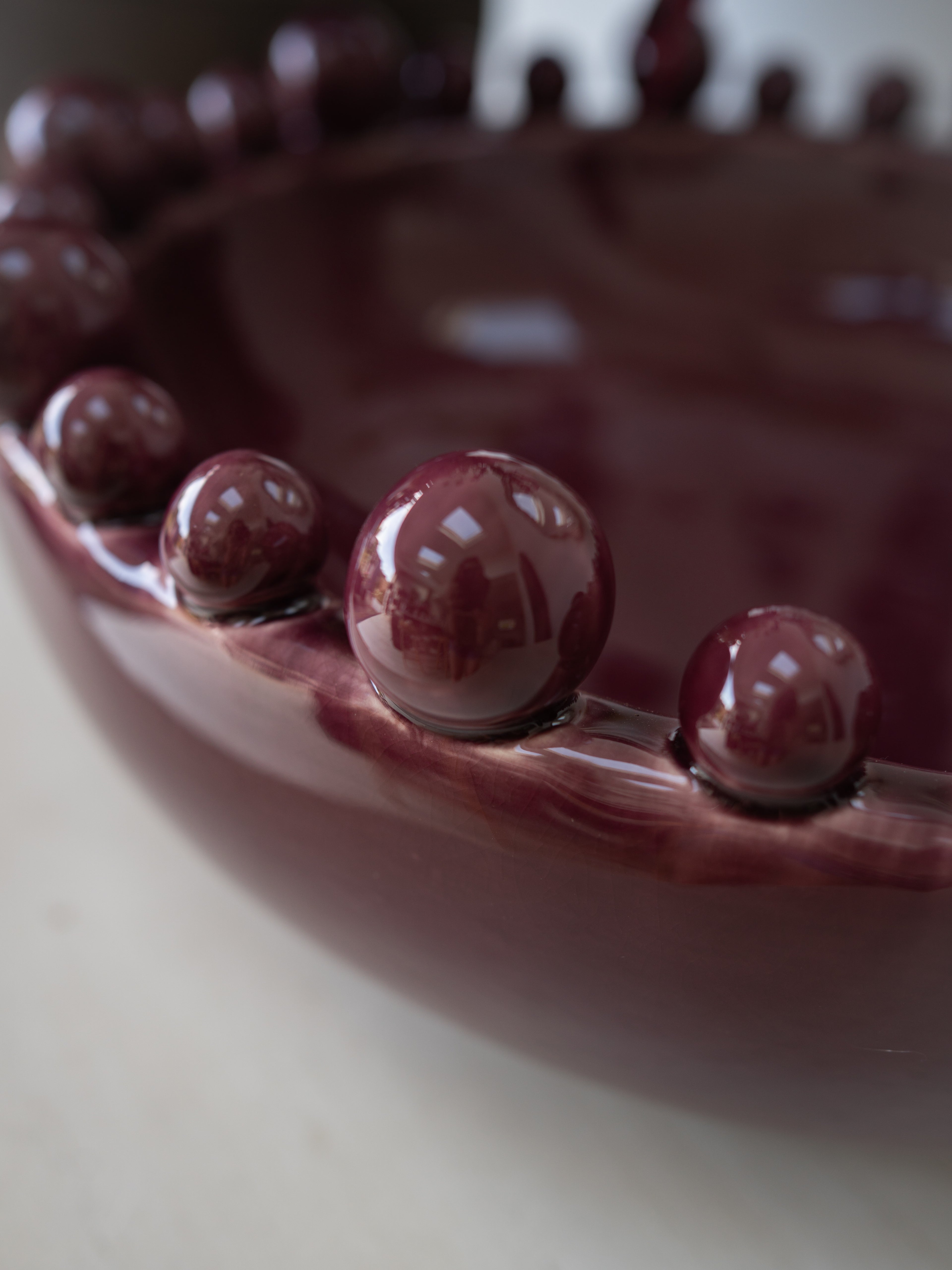 Close-up of a glossy dark burgundy bowl with a decorative border of raised round beads, photographed on a light neutral surface.