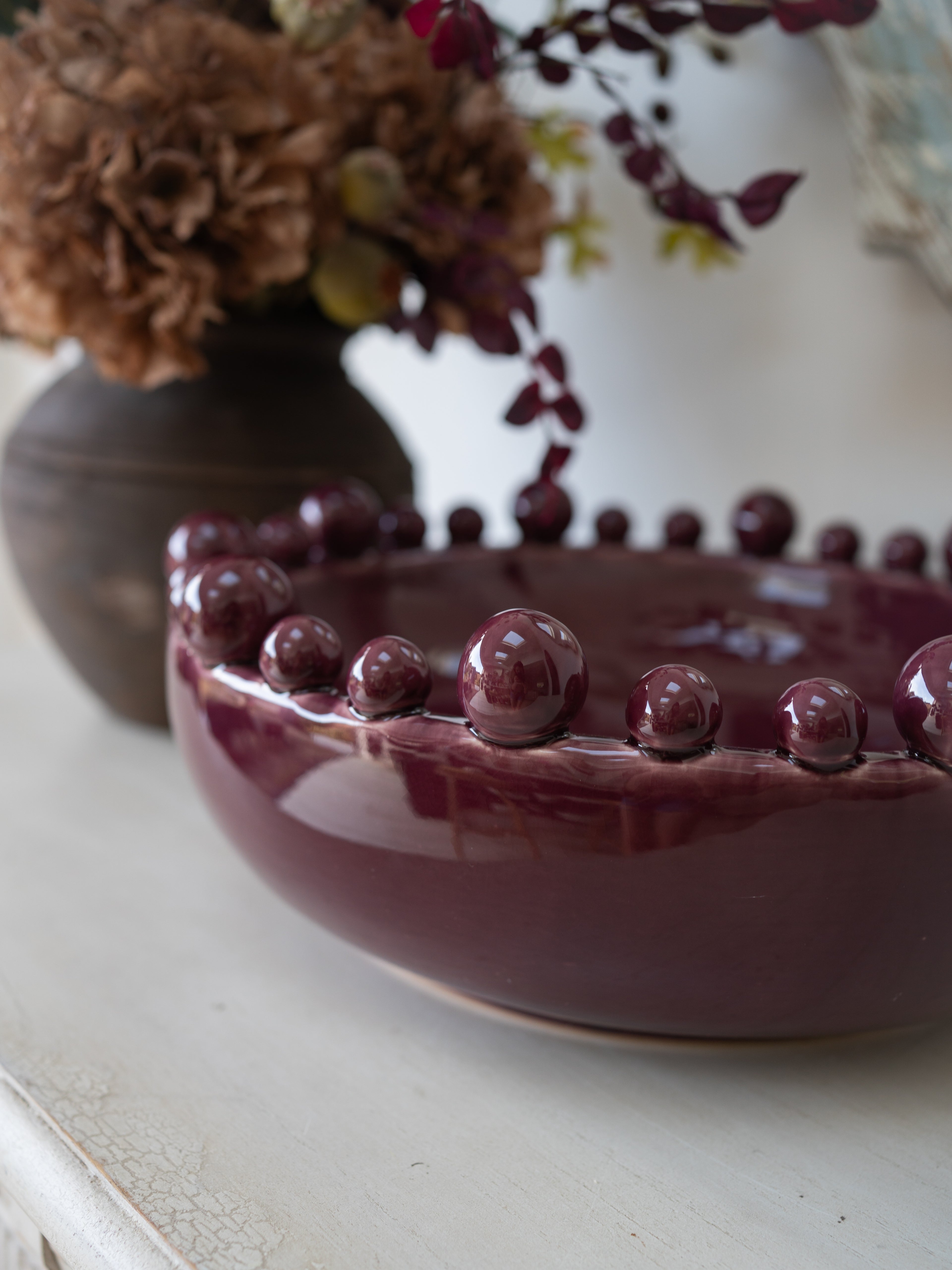 A glossy deep burgundy ceramic bowl with a beaded rim, styled on a white surface with a blurred dark vase of dried flowers in the background.