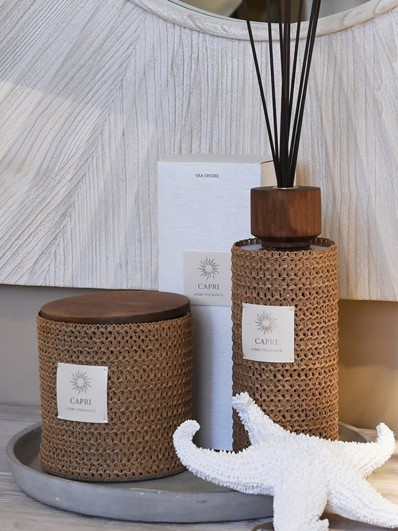 Two woven reed diffuser and candle with capri labels on a surface with a textured wall in the background