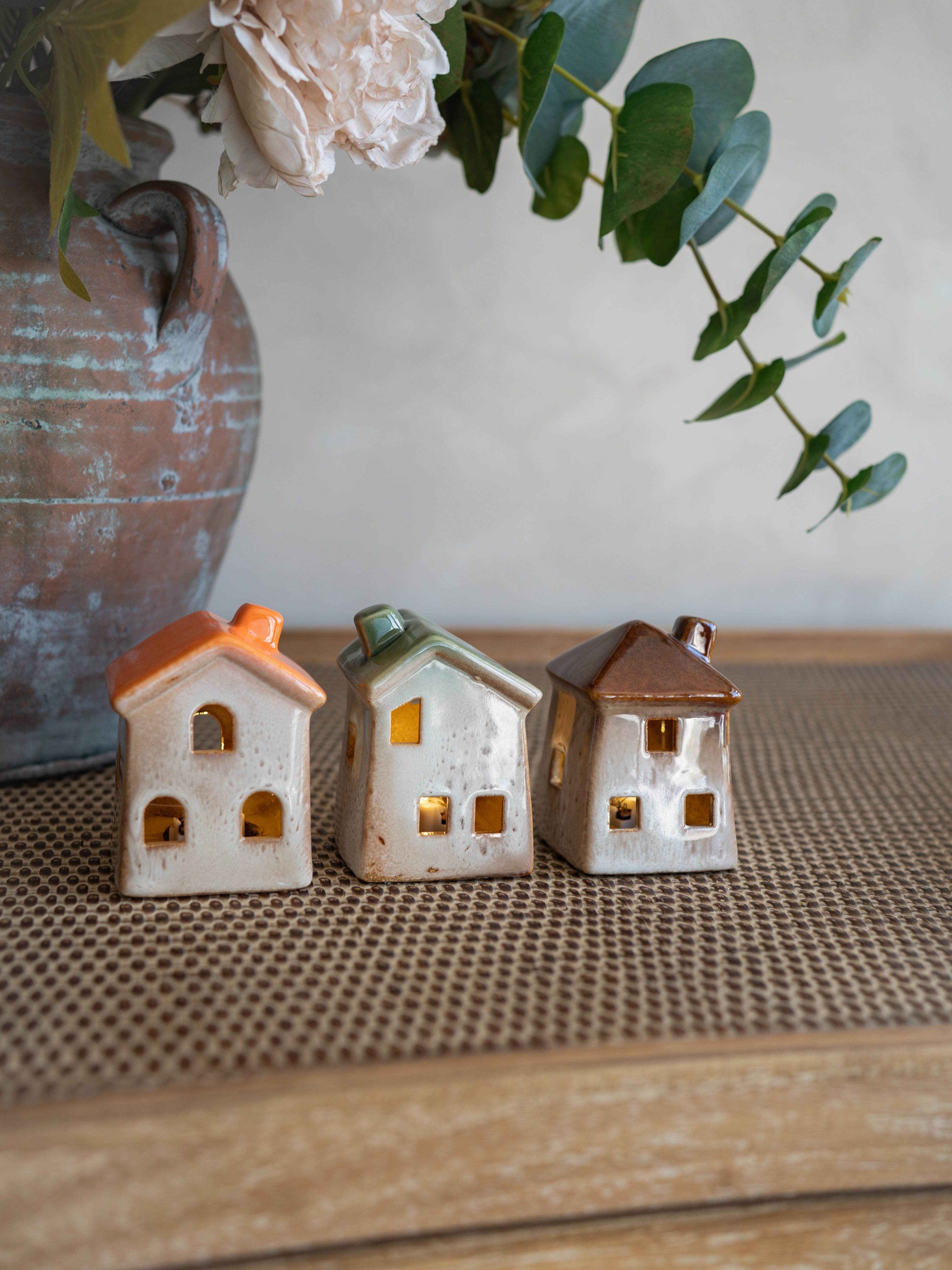 Three small ceramic house-shaped ornaments with orange, green, and brown roofs, displayed on a woven brown mat with a rustic vase and greenery in the background.