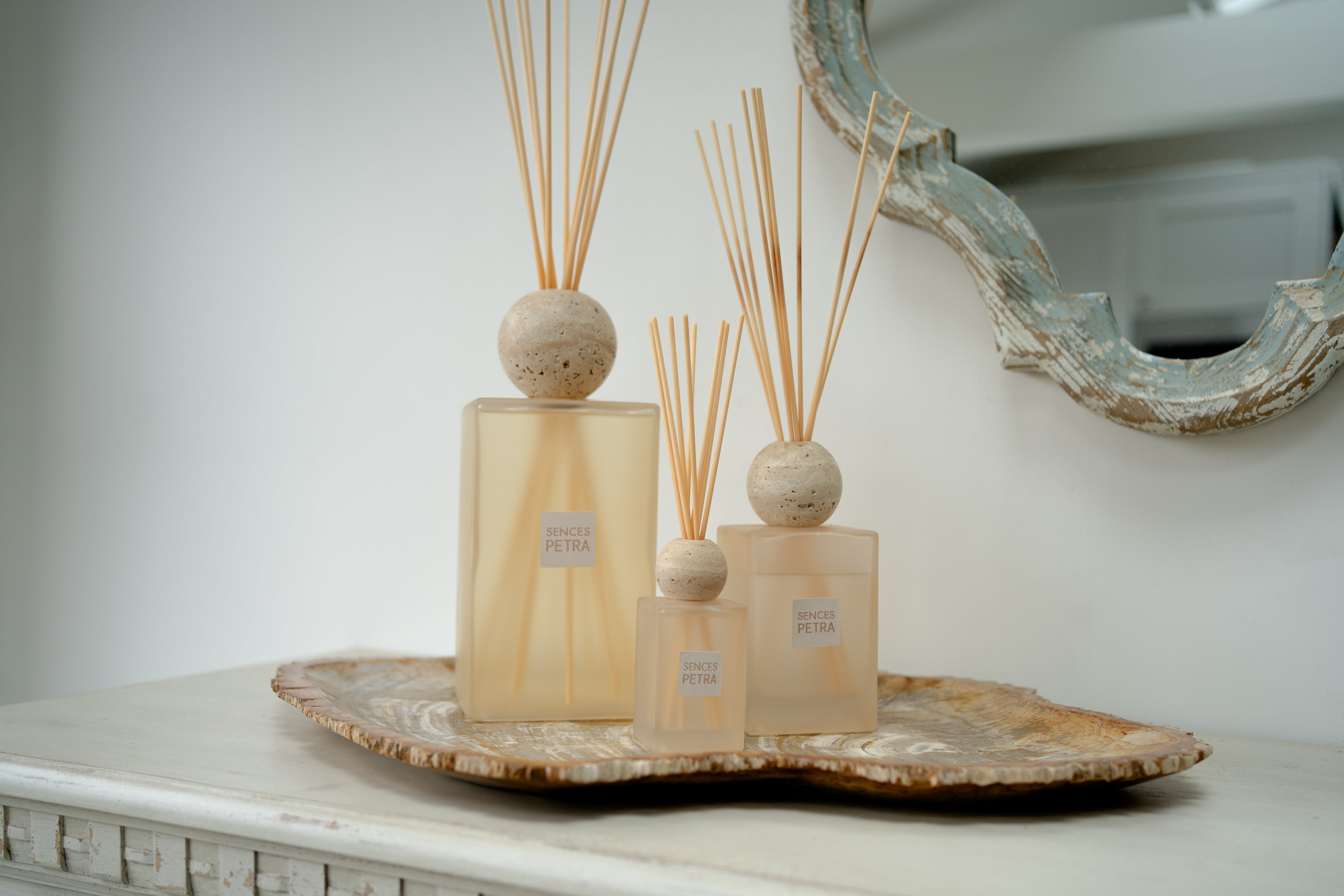 Three petra sences reed diffusers with frosted beige glass bottles and natural wood reeds, arranged on a decorative tray against a soft white wall.