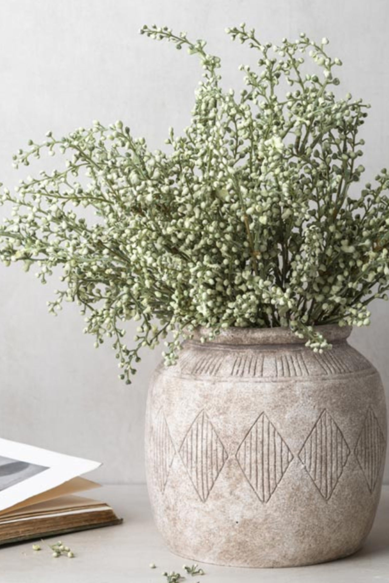 A textured beige ceramic vase with a carved diamond pattern, filled with faux green heather stem, set against a neutral light gray background.
