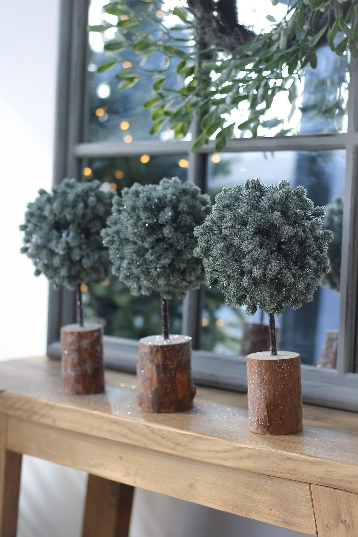 Three frosted green topiary christmas trees in rustic brown log planters, arranged on a light wood table in front of a window.