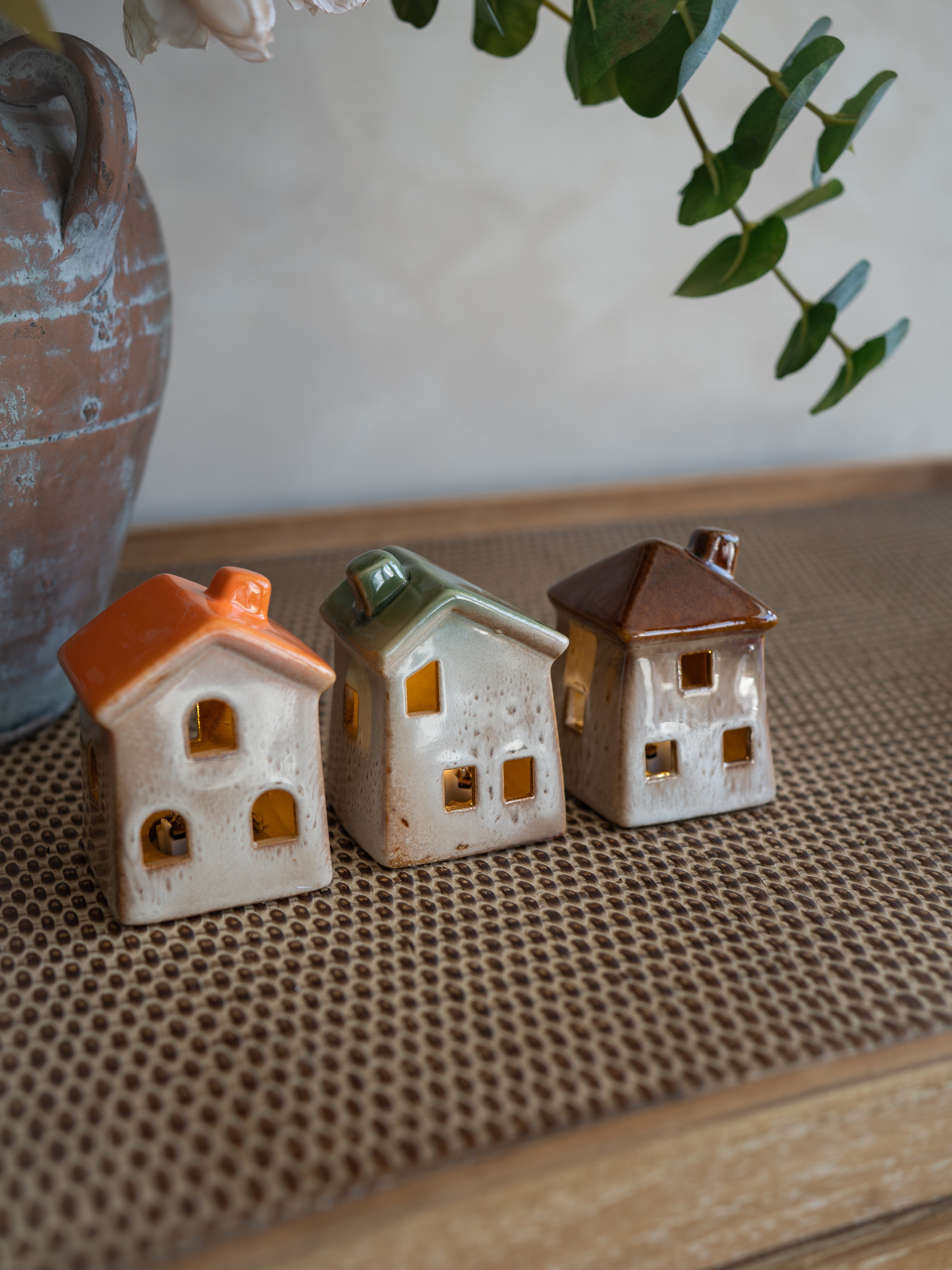 Three small ceramic house-shaped ornaments with orange, green, and brown roofs, displayed on a woven brown mat with a rustic vase and greenery in the background.