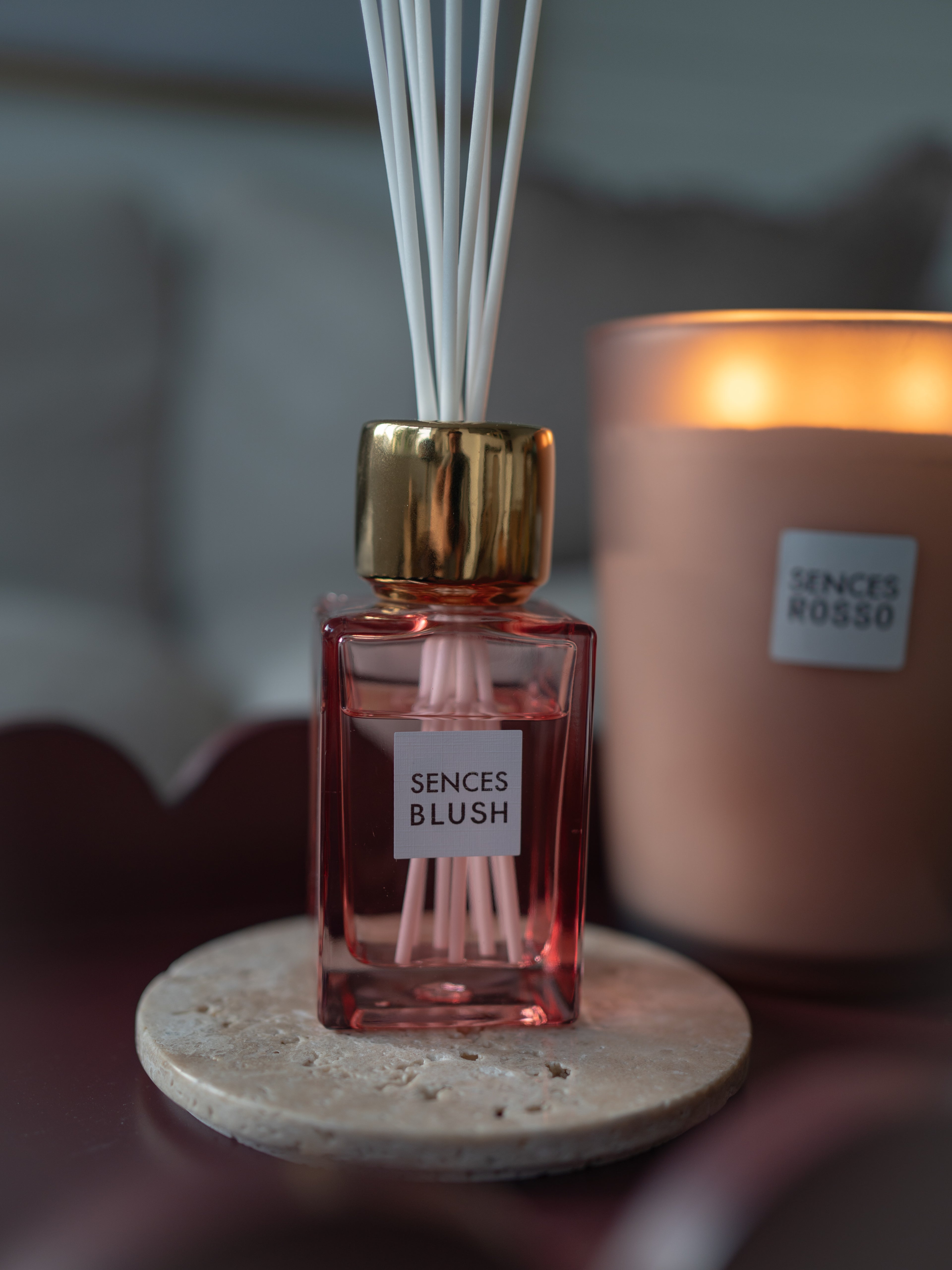 A Senses Blush reed diffuser bottle with pink liquid, white reeds, and a gold cap sits on a stone coaster in the foreground, with a blurred Senses Rosso candle in the background.