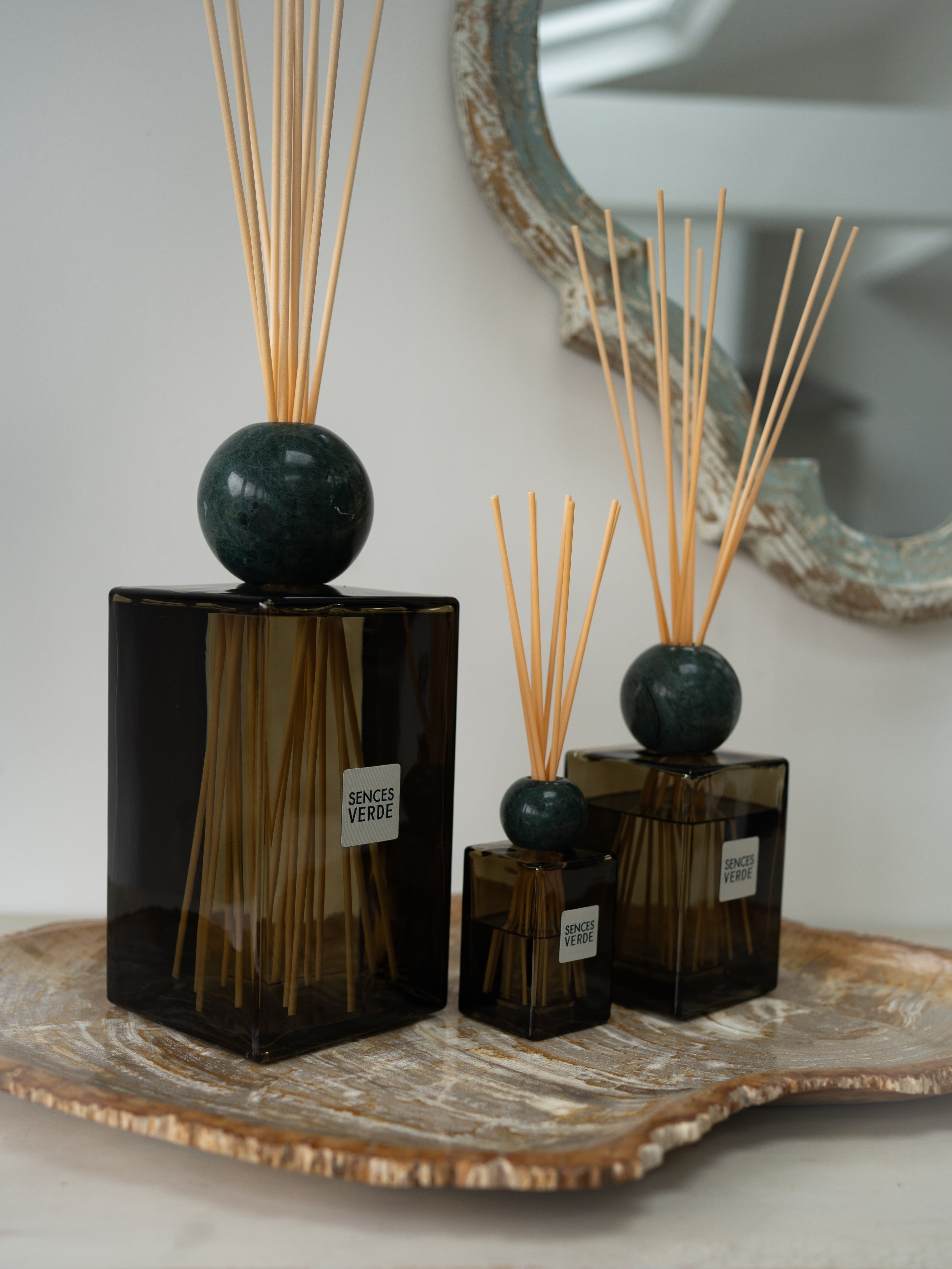 Three reed diffusers in dark green glass bottles with round dark green stone tops and natural reed sticks, arranged on a decorative tray against a soft neutral background.