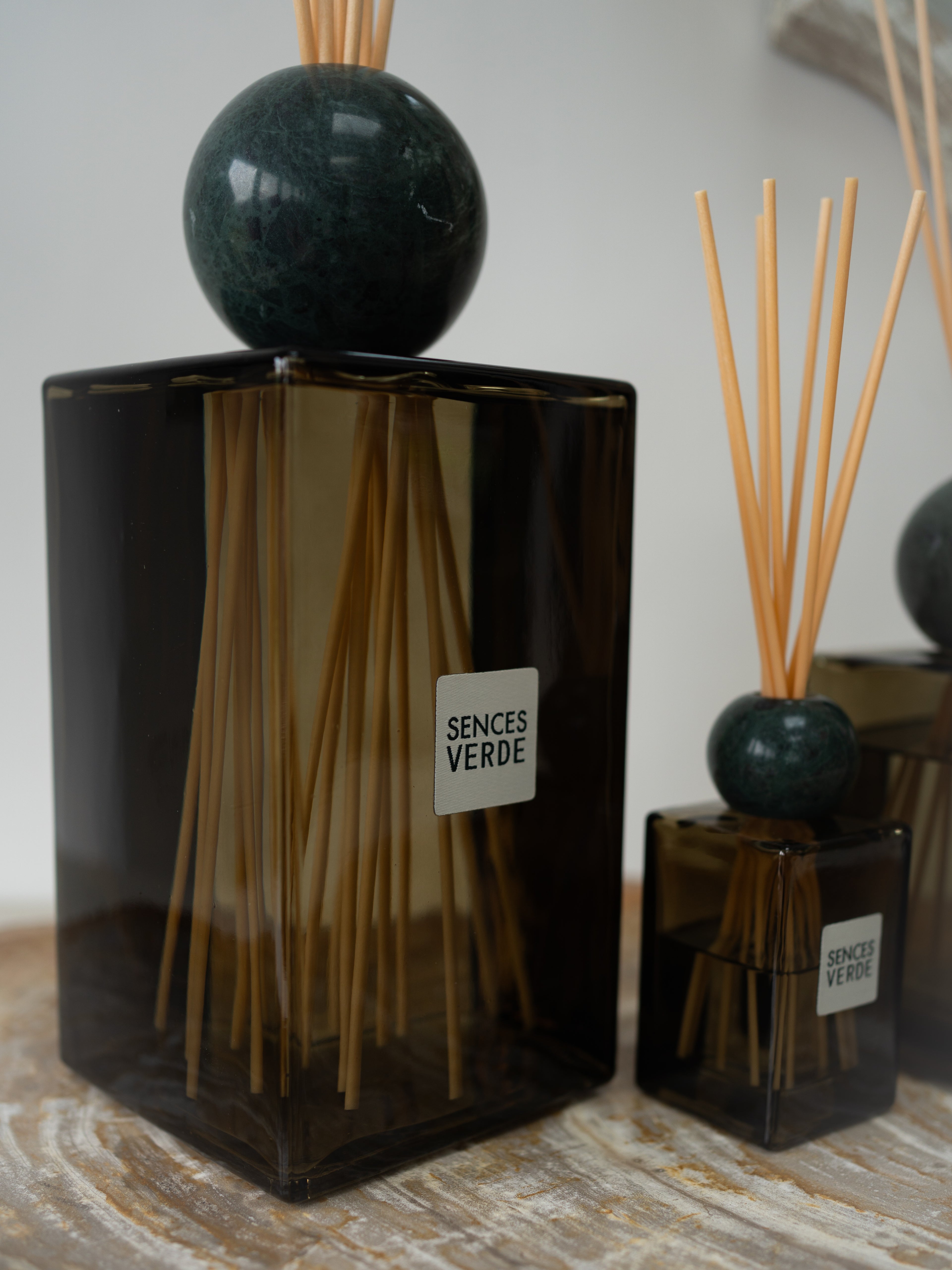 A Sences Verde reed diffuser set featuring a large green glass bottle with a dark green marble sphere and wooden reeds, alongside a smaller matching bottle on a rustic wooden surface.