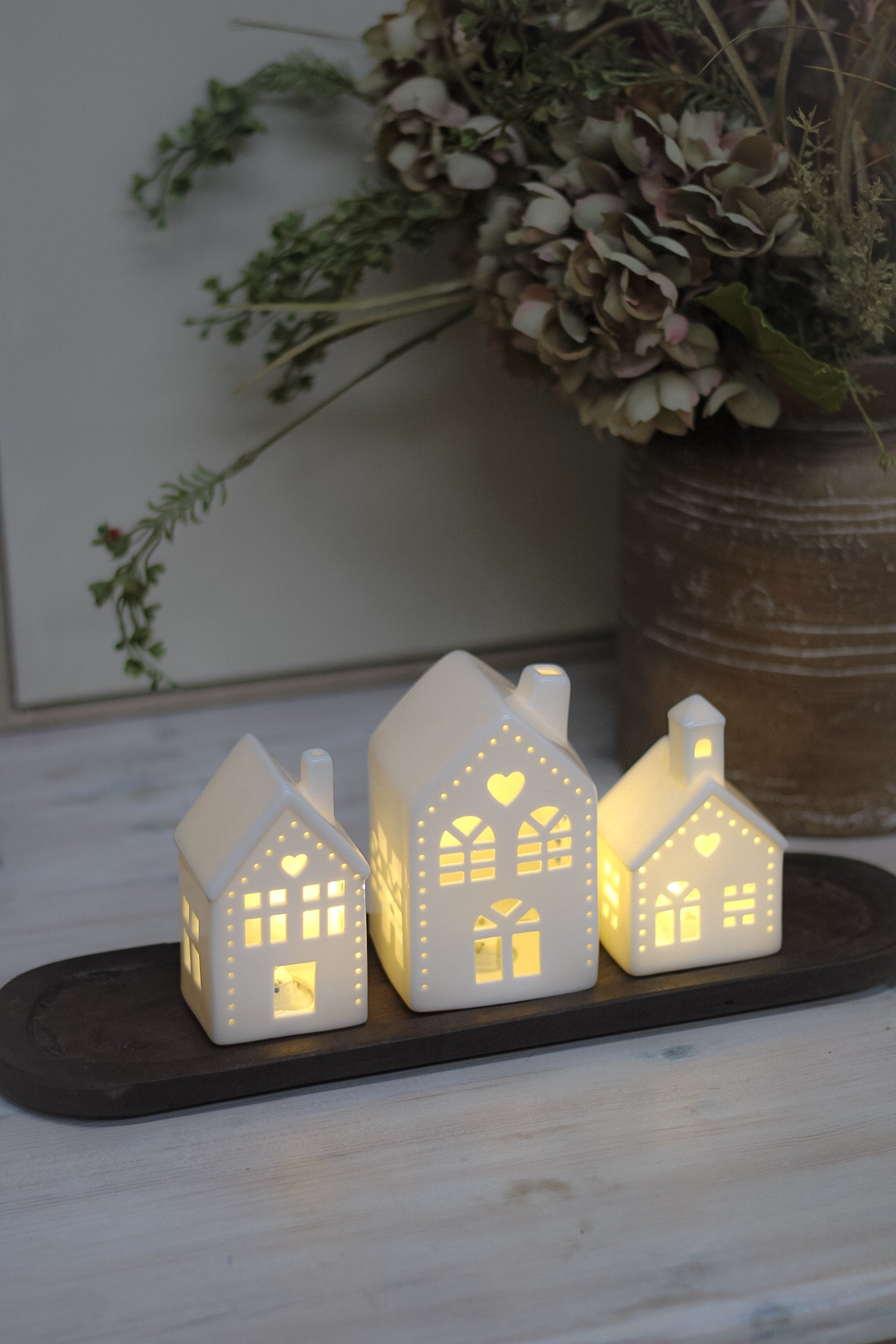 White ceramic houses with cut-out hearts and dots design, illuminated from the inside by LED lights, placed on a wooden surface.