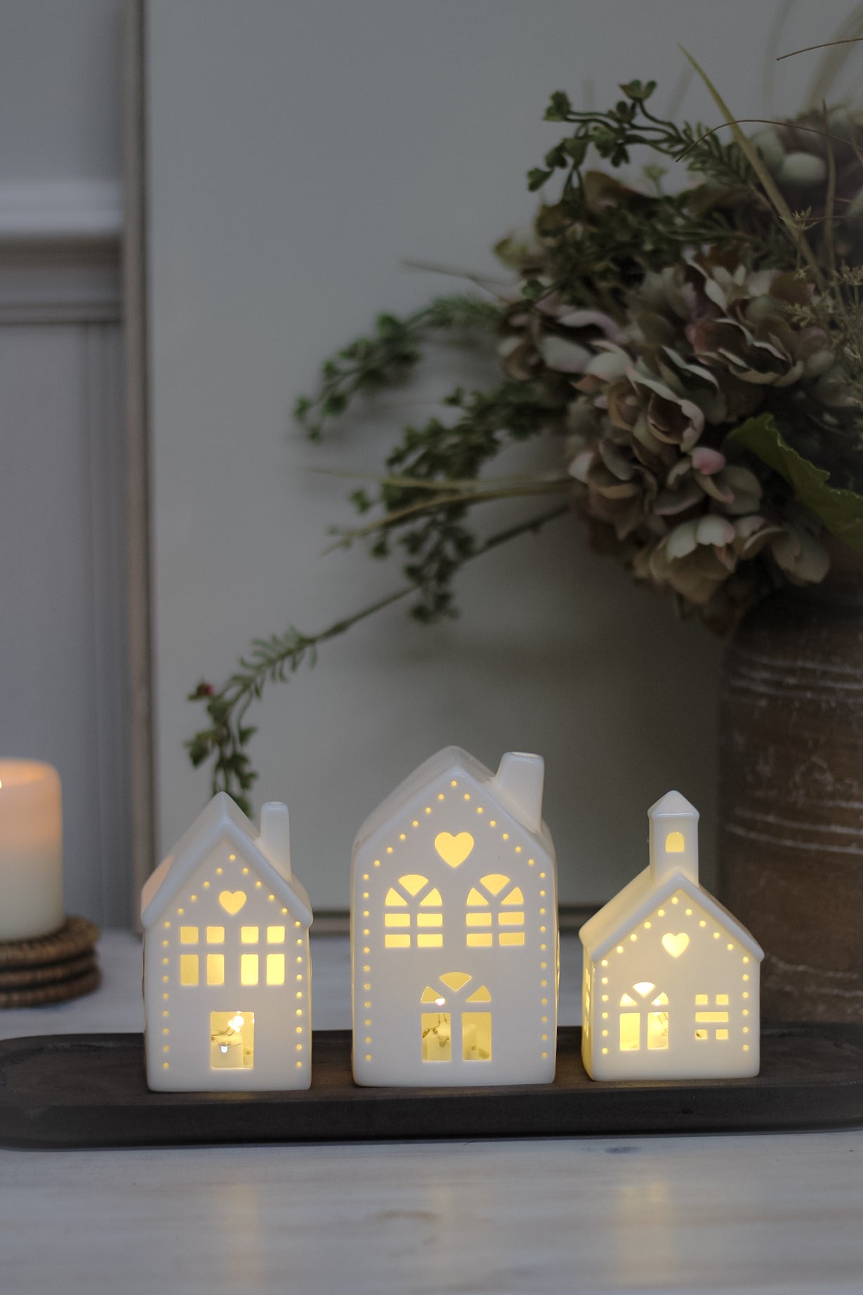 White ceramic houses with cut-out hearts and dots design, illuminated from the inside by LED lights, placed on a wooden surface.