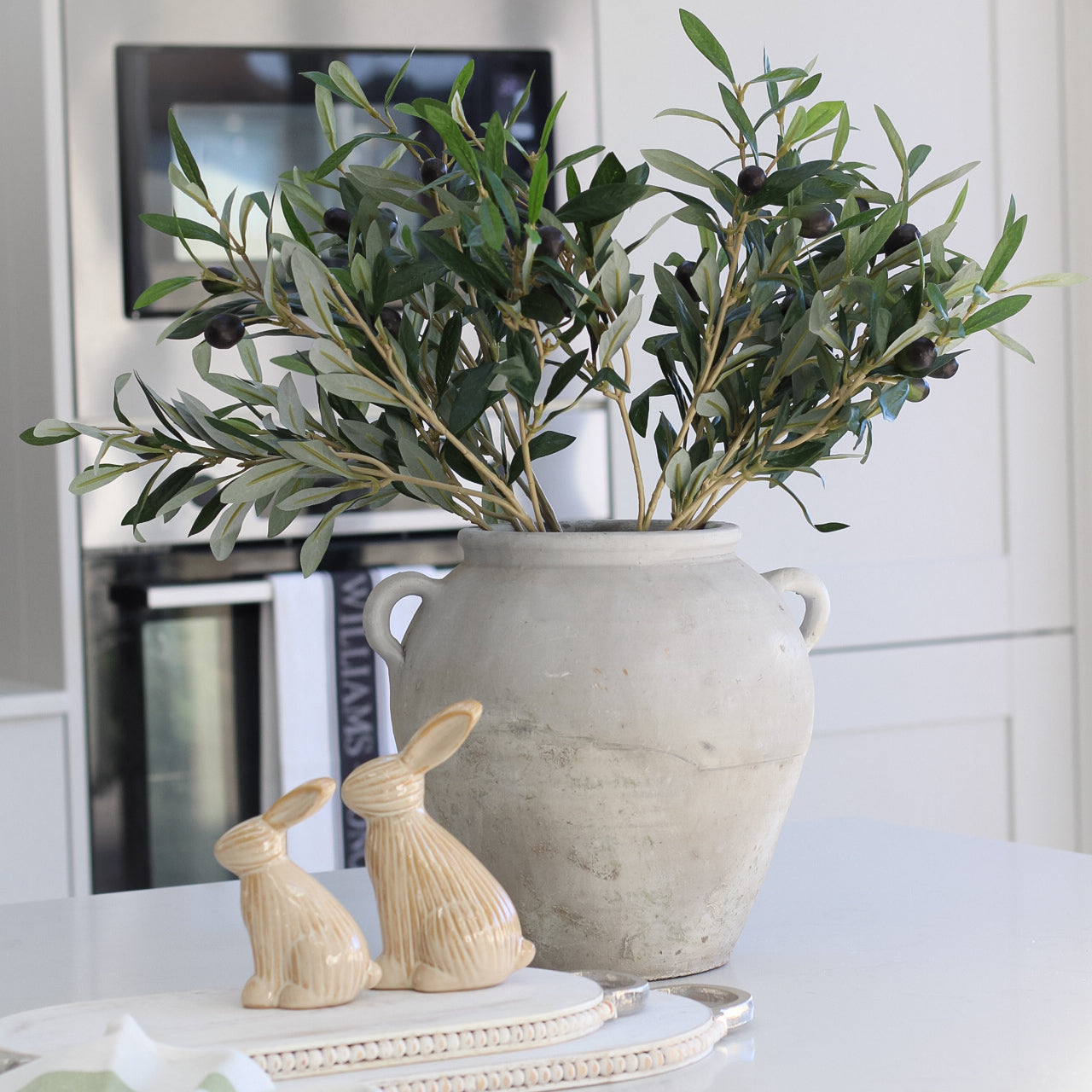 Tall grey olive pot, filled with faux olive stems, next white wooden beaded board with pair of natural toned bunnies with line pattern