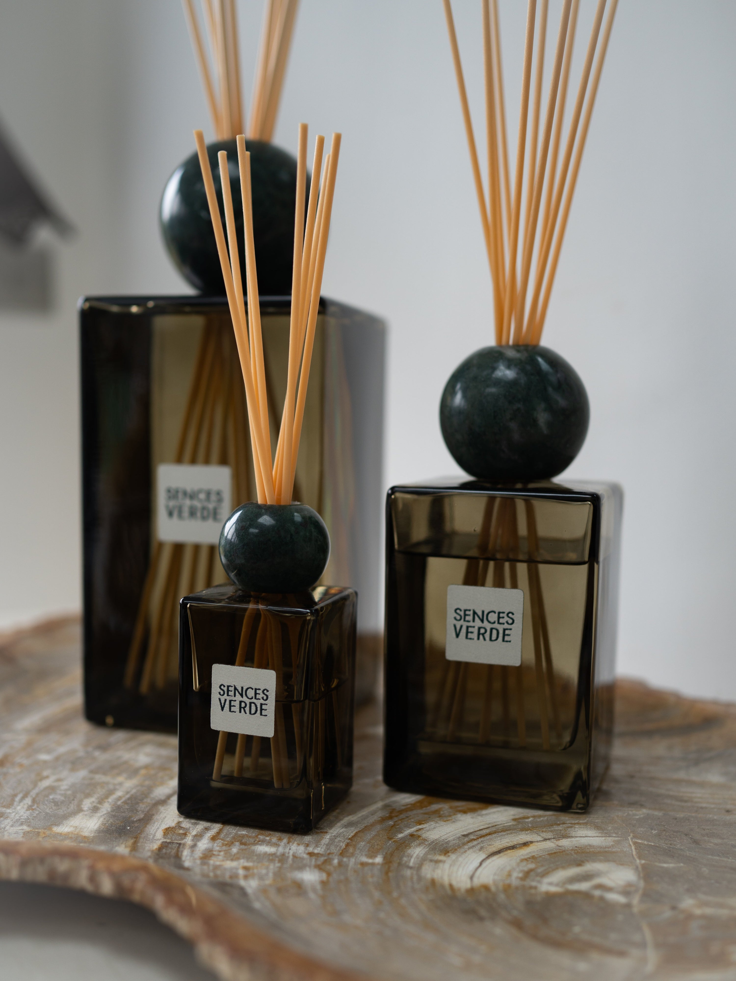 Three Sences Verde reed diffusers in varying sizes, featuring dark amber glass bottles, round black stone tops, and natural wood reeds, arranged on a fossilled wood slice tray.