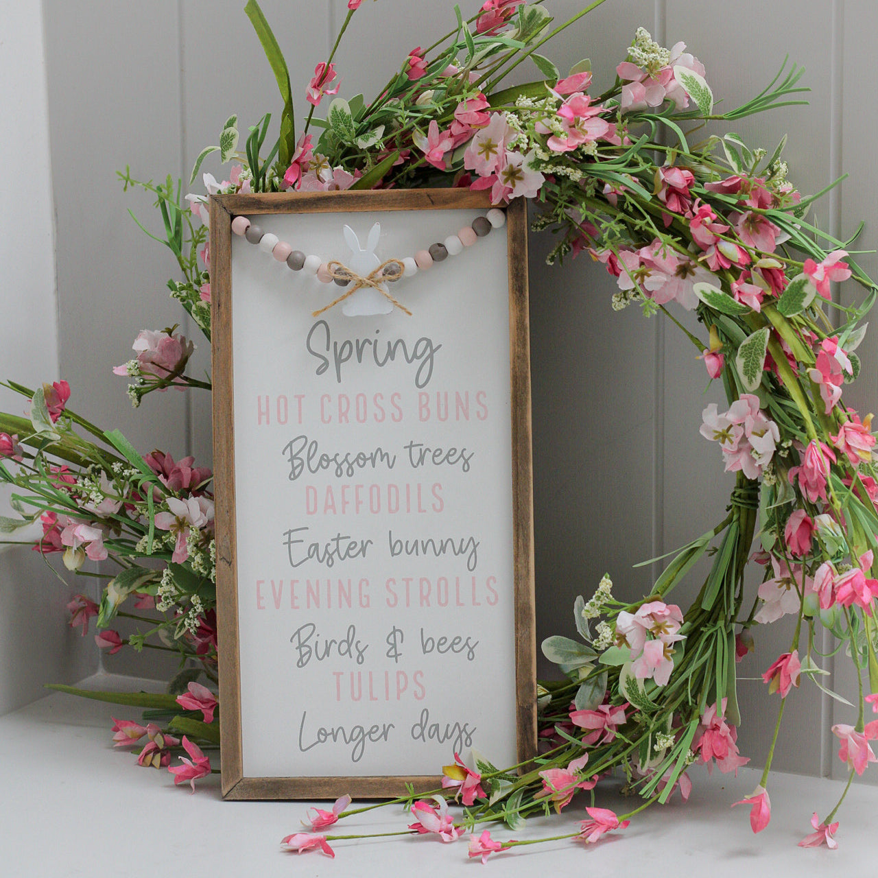 Rustic framed wooden plaque with white background with beaded hanging bunny detail and list of favourite things about Spring 'hot cross buns, Blossom trees, Daffodils, Easter bunny, Evening strolls, Birds and bees, tulips and longer days' with pink floral wreath behind