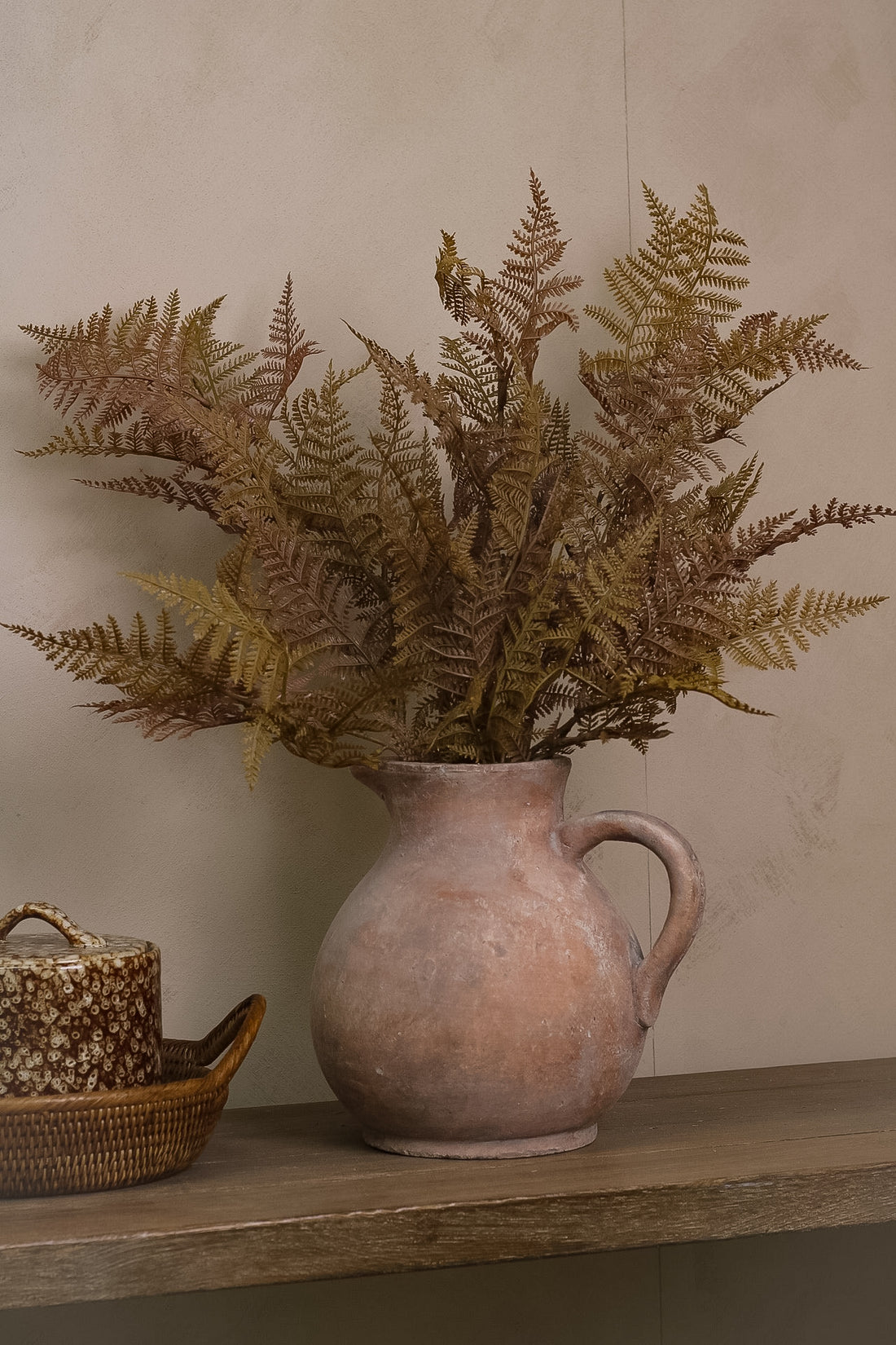 Faux ferns in a terracotta pitcher on a wooden shelf against a beige wall