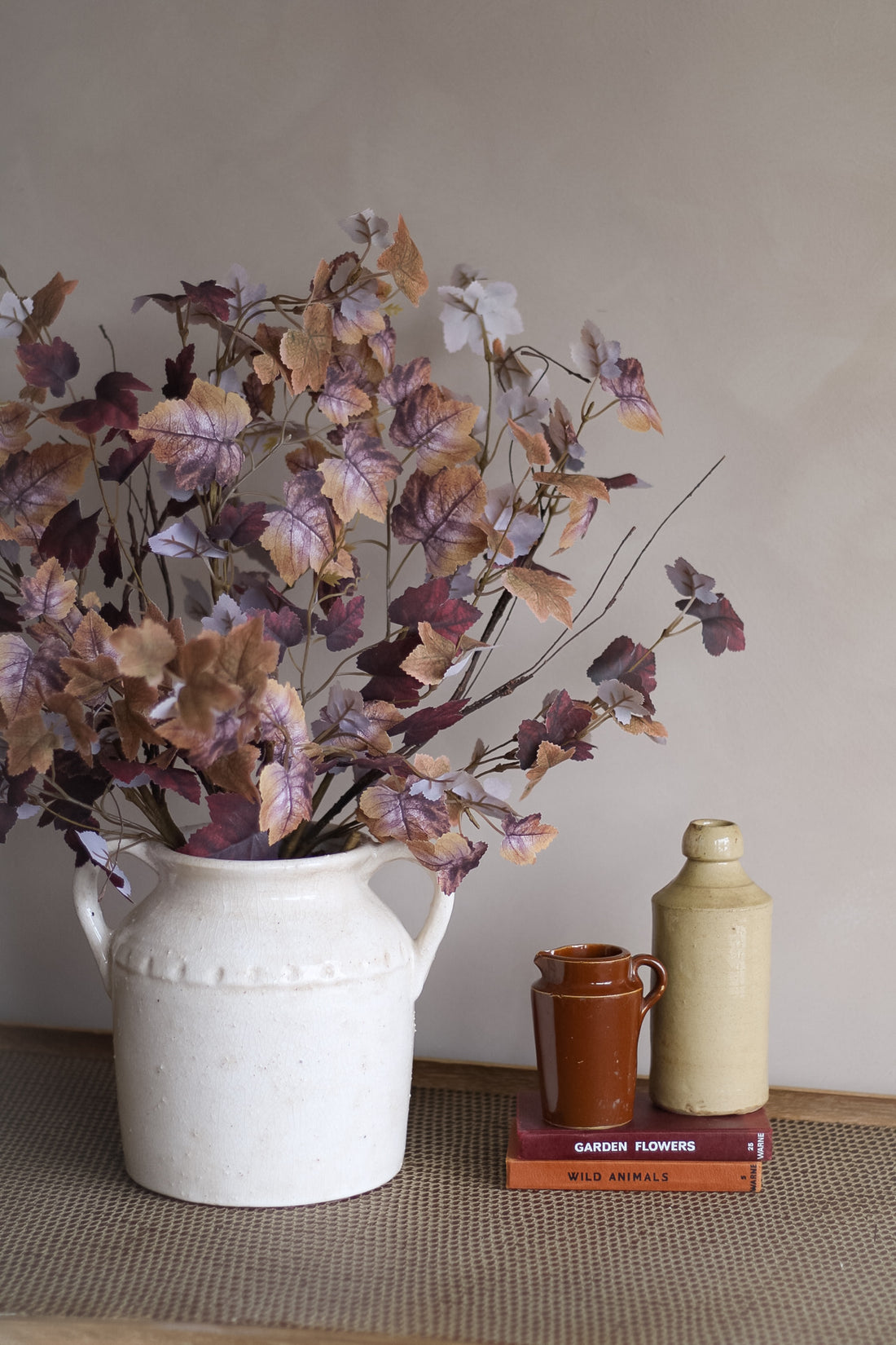 Decorative faux rustic leaves arrangement with a white vase, brown vintage jug, cream bottle, and books on a textured surface.